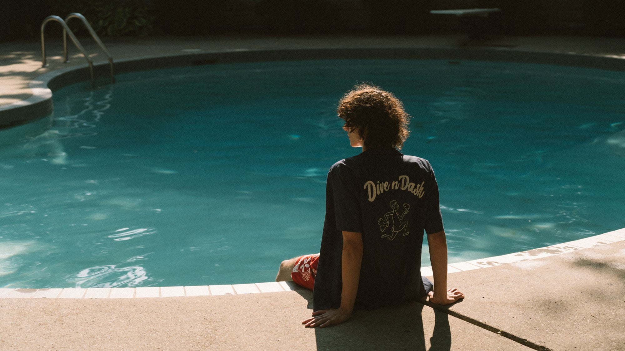 A model sitting by the pool wearing Bather's navy embroidered Dive n' Dash Camp Shirt
