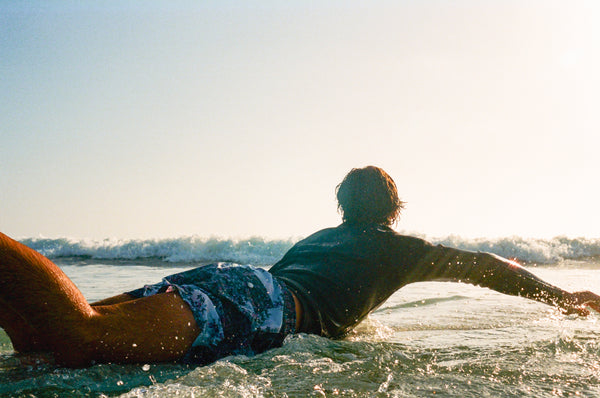 Bather model on surfboard in the ocean wearing pepperbush swim trunks