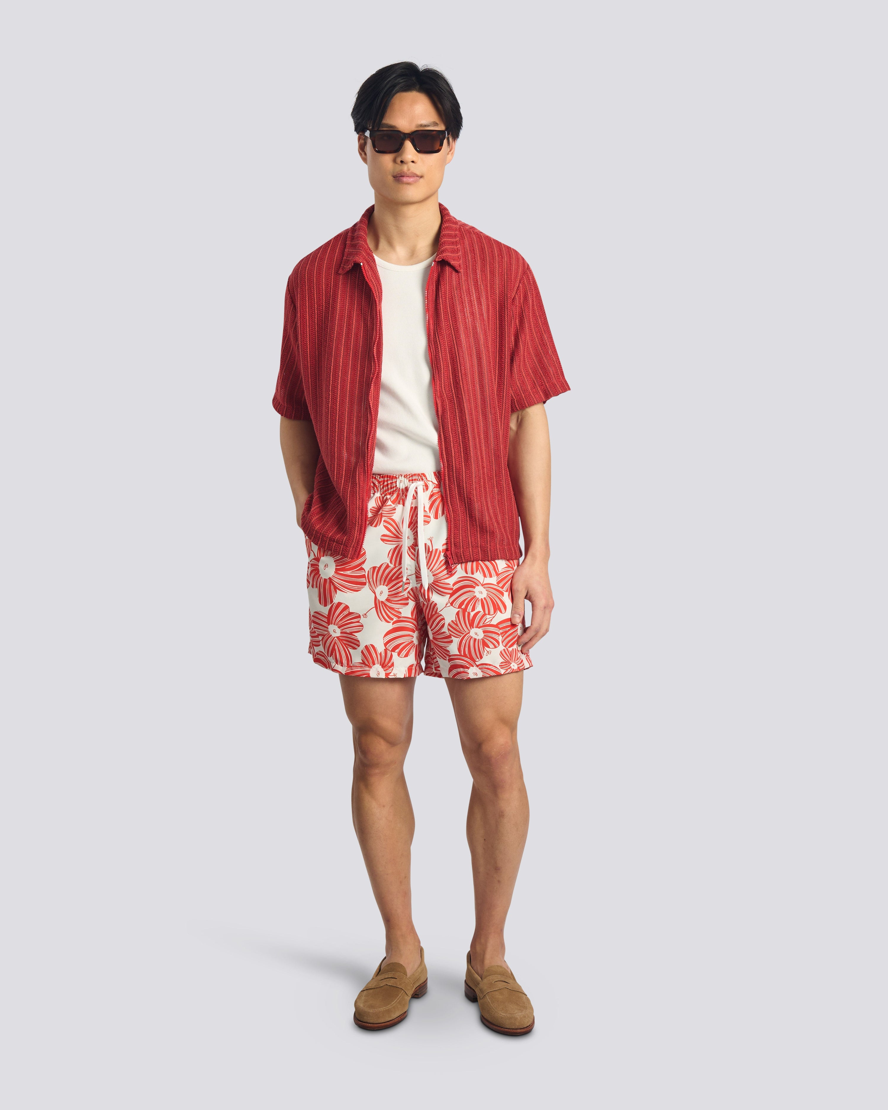 Person wearing a red shirt, white and red patterned shorts, and brown shoes on a light gray background