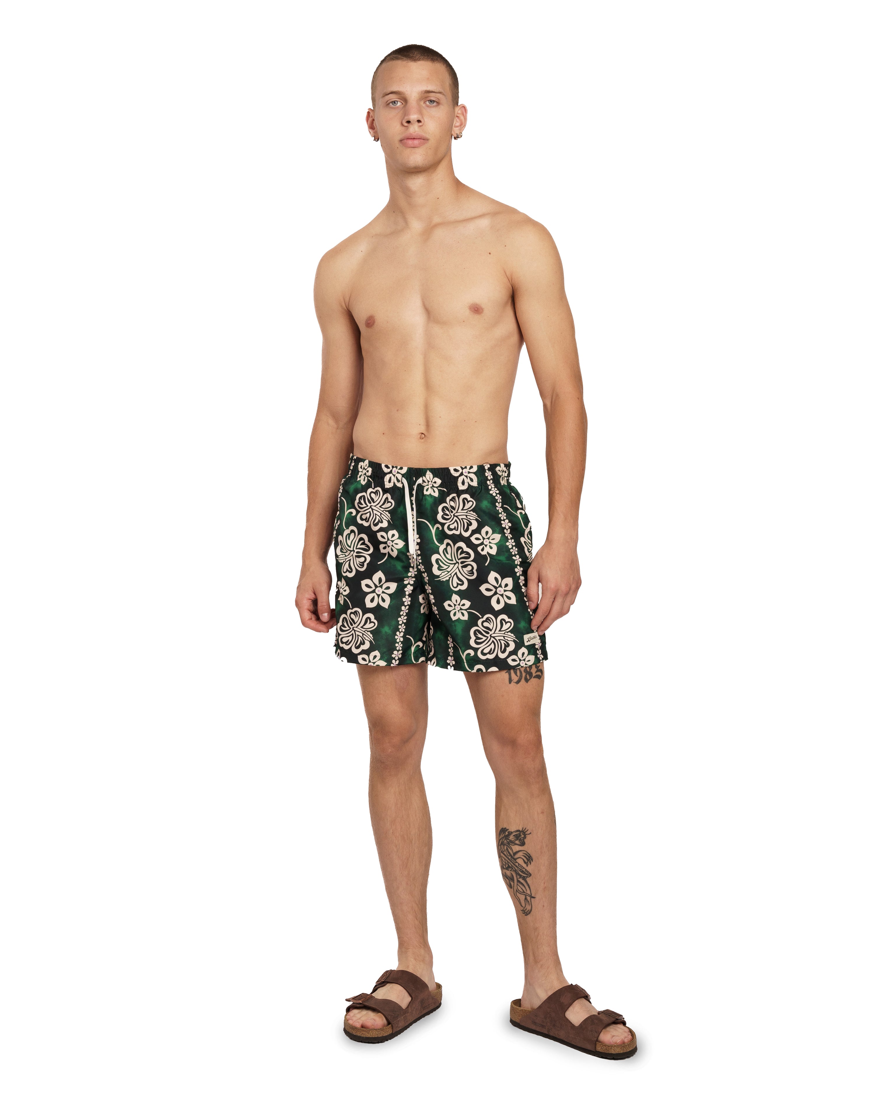 Model wearing Green Bather swim trunk with a tropical pattern and beige floral motif
