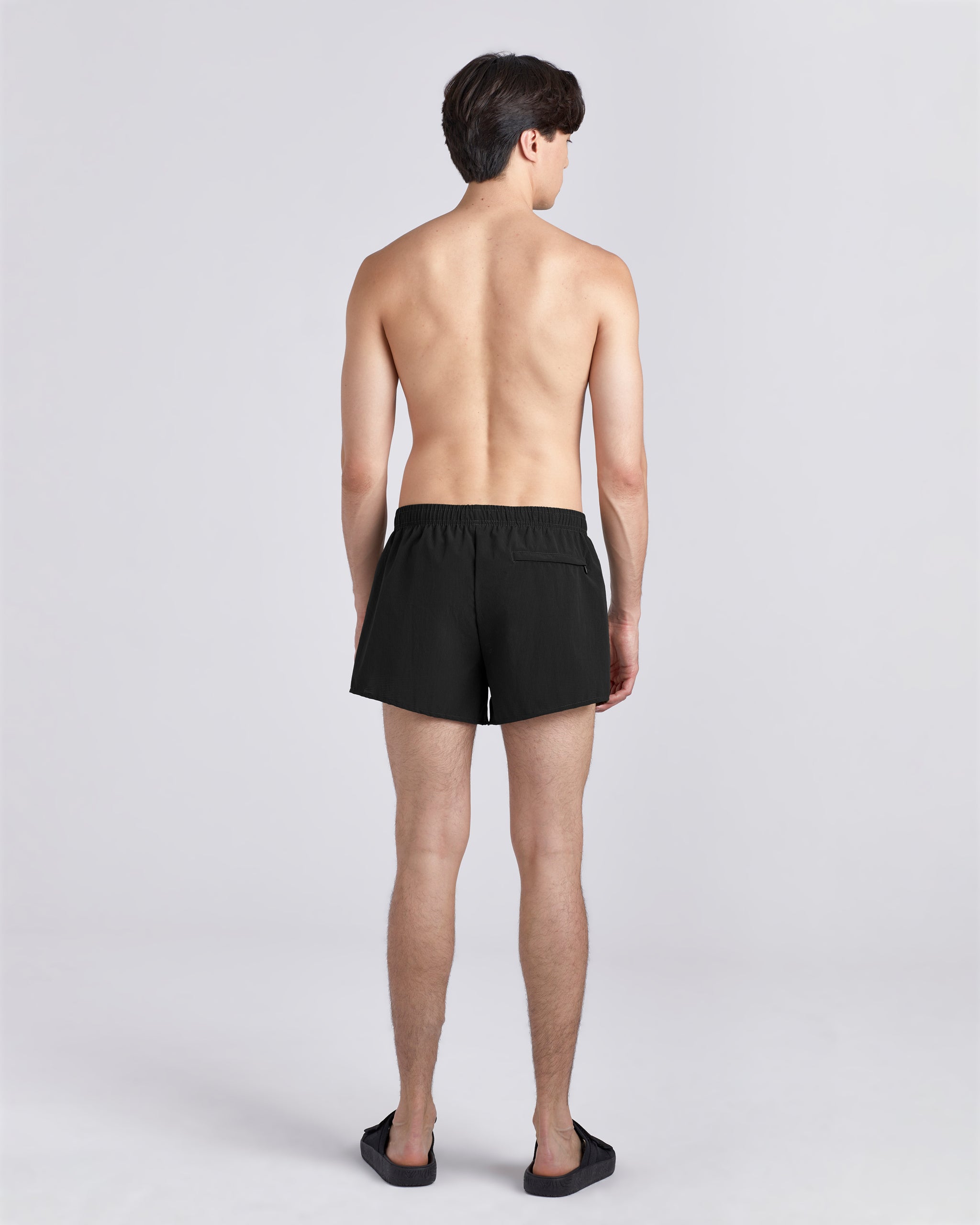 Back view of shirtless model wearing sandals and 3.5 inch inseam Bather Active Trunk in black featuring lightweight recycled Japanese DotAir nylon with stretch fabric and compression liner