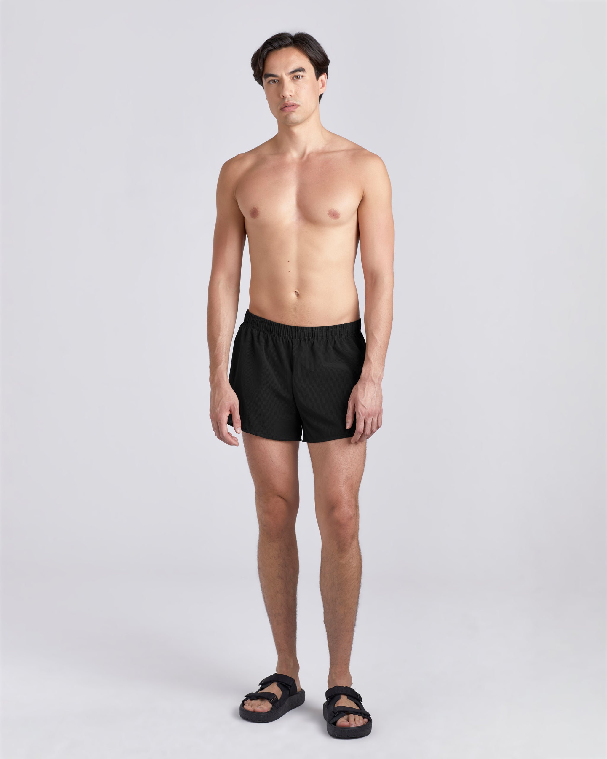 Front view of shirtless model wearing sandals and 3.5 inch inseam Bather Active Trunk in black featuring lightweight recycled Japanese DotAir nylon with stretch fabric and compression liner