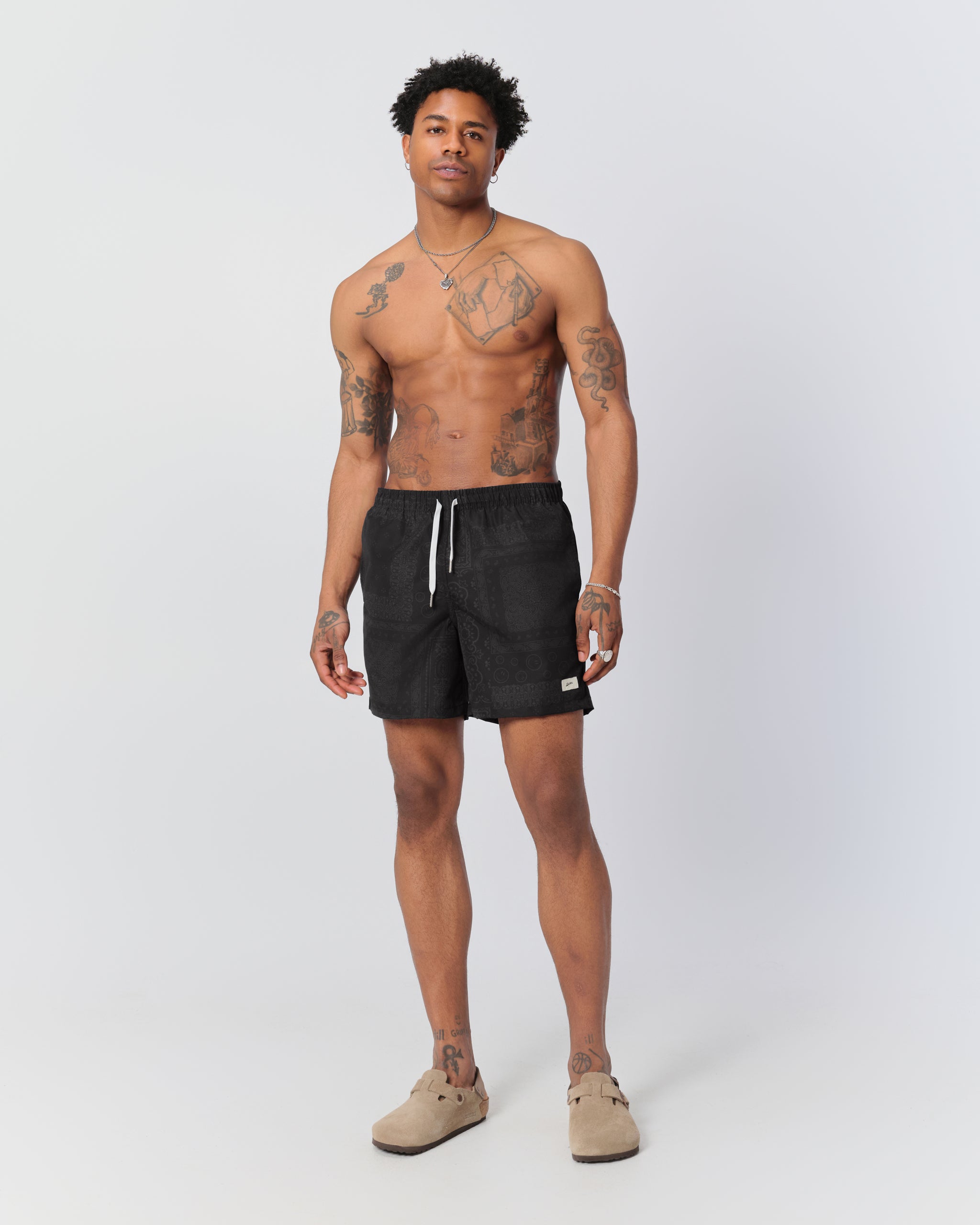 Man wearing Bather black swim trunks with bandana pattern, elastic waistband, and white drawstring