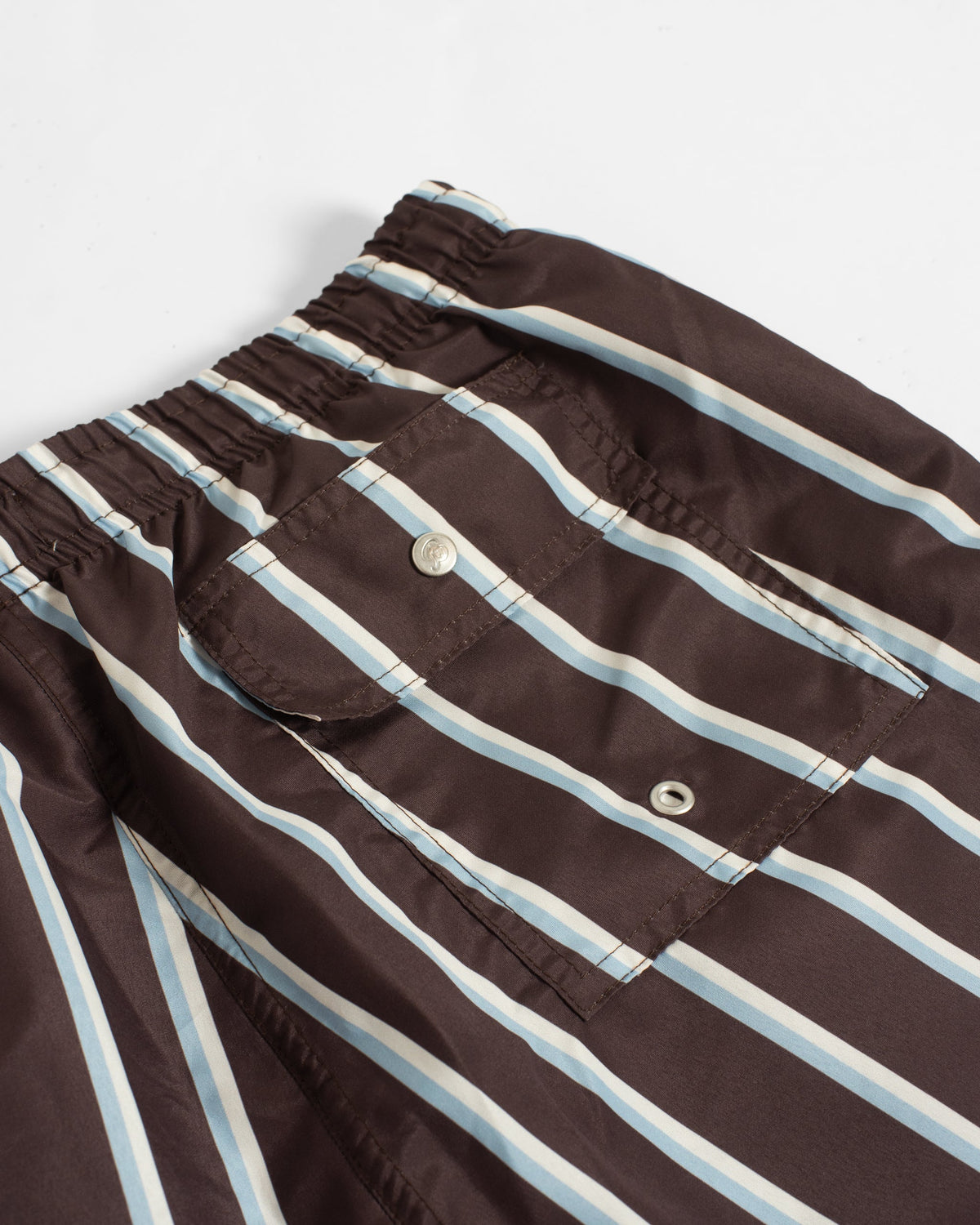 Close-up of a brown striped garment, likely a jacket or overshirt, featuring vertical stripes in light blue and off-white, with a buttoned chest pocket and elastic waistband.