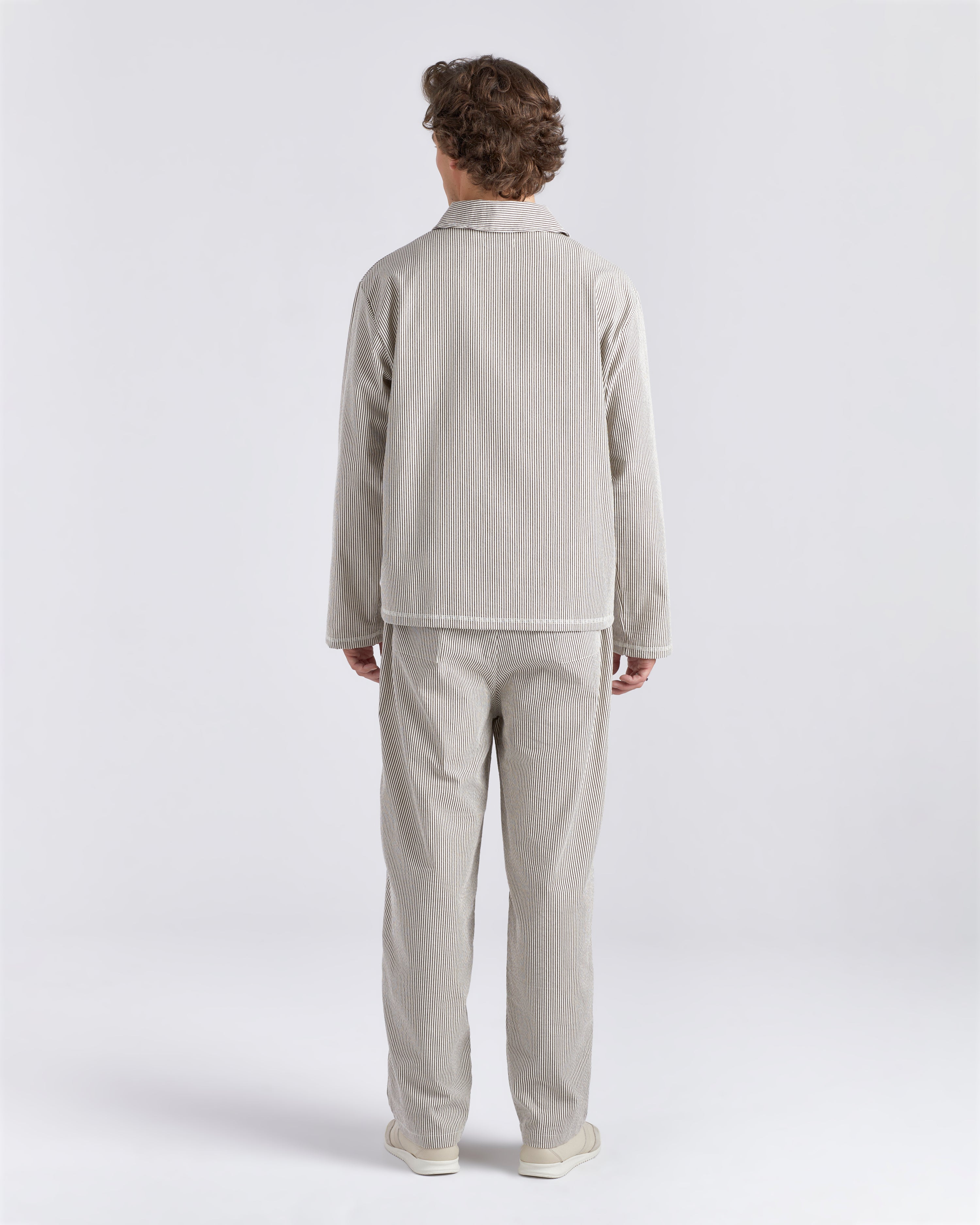 Back of male model wearing a Bather brown pinstripe tour jacket featuring a front zipper with matching brown Bather pin strip pants.