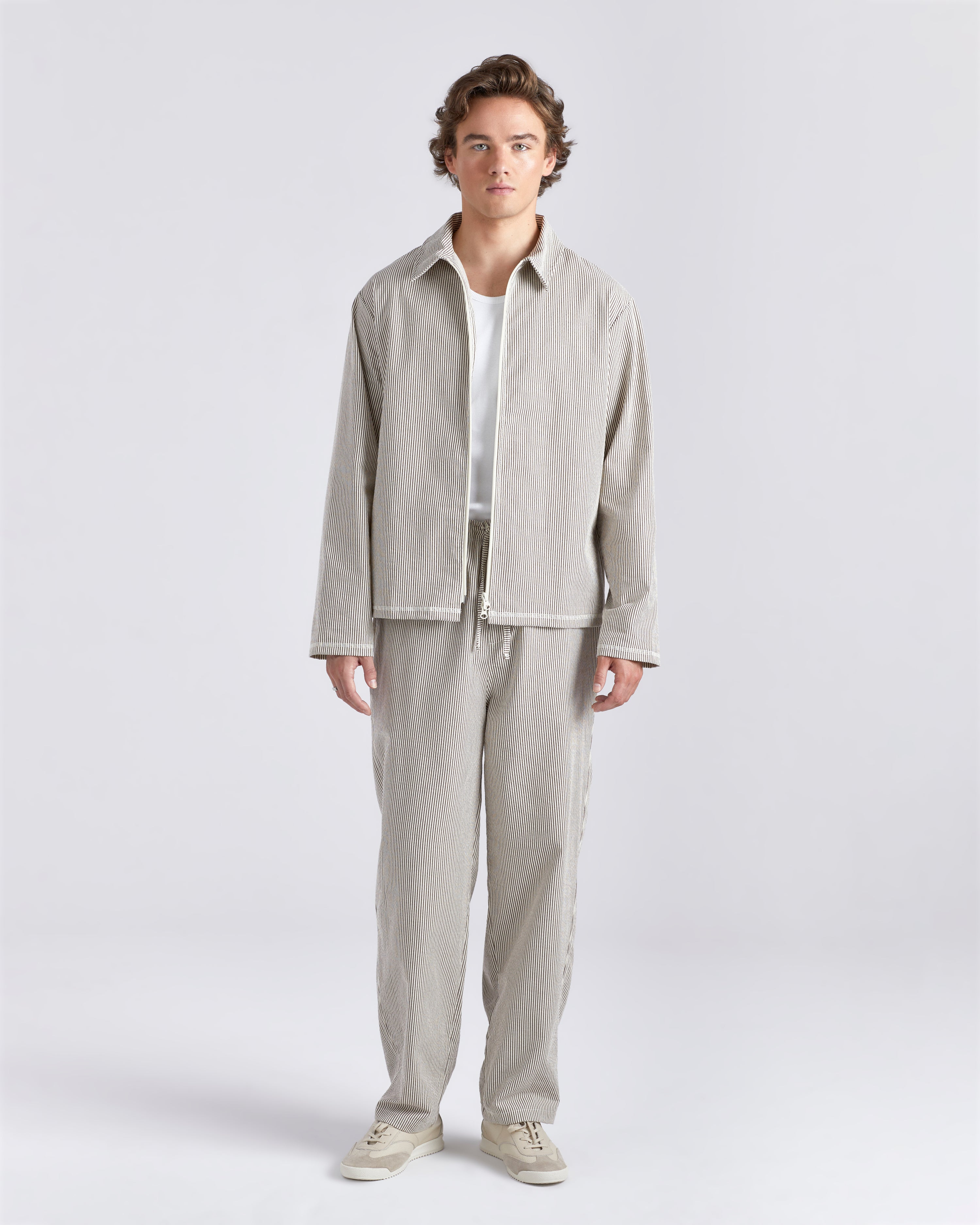 Male model wearing a Bather brown pinstripe tour jacket featuring a front zipper with matching brown Bather pin strip pants.