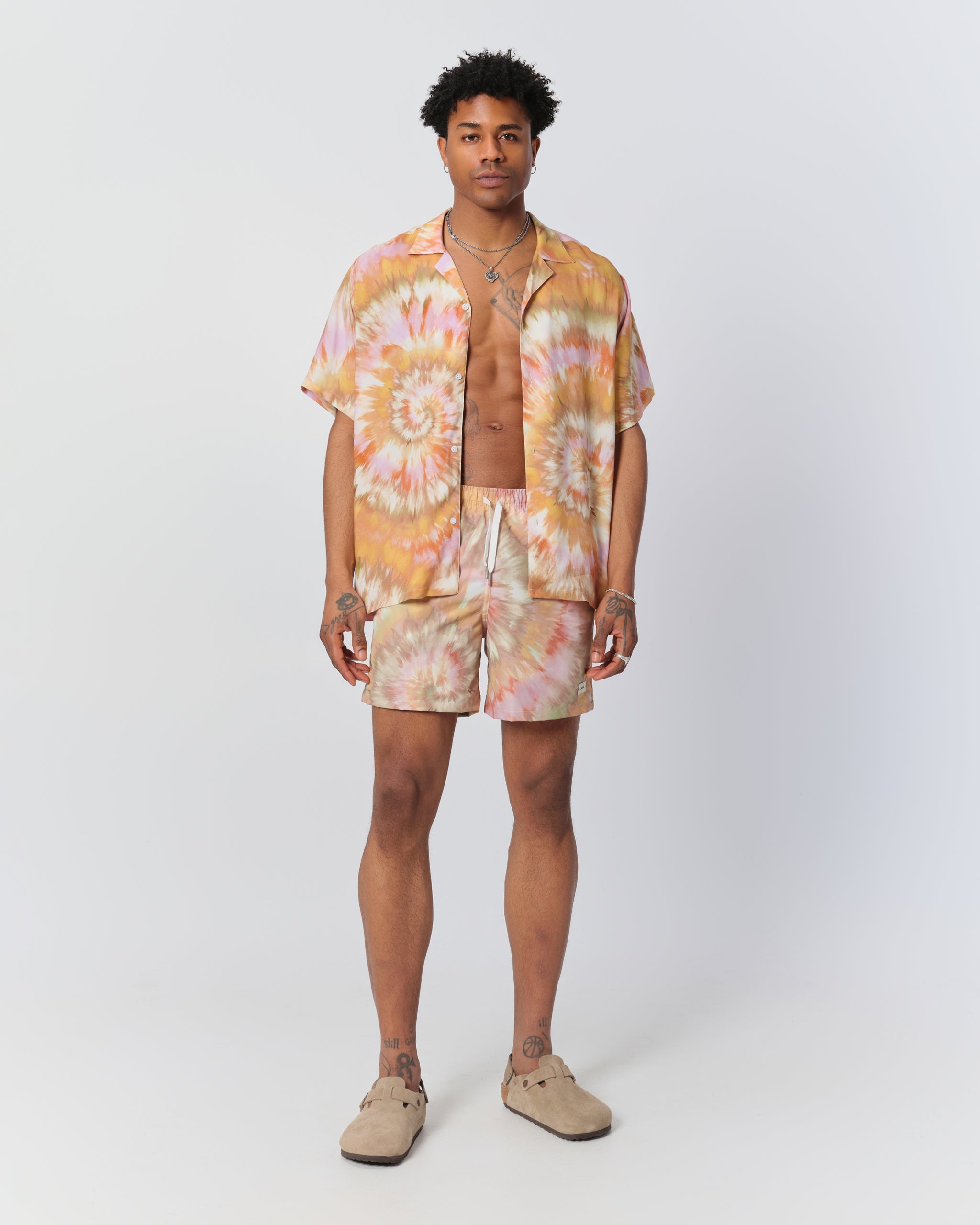Man wearing abstract tie-dye swim trunks in warm orange and peach tones and matching Bather camp shirt 