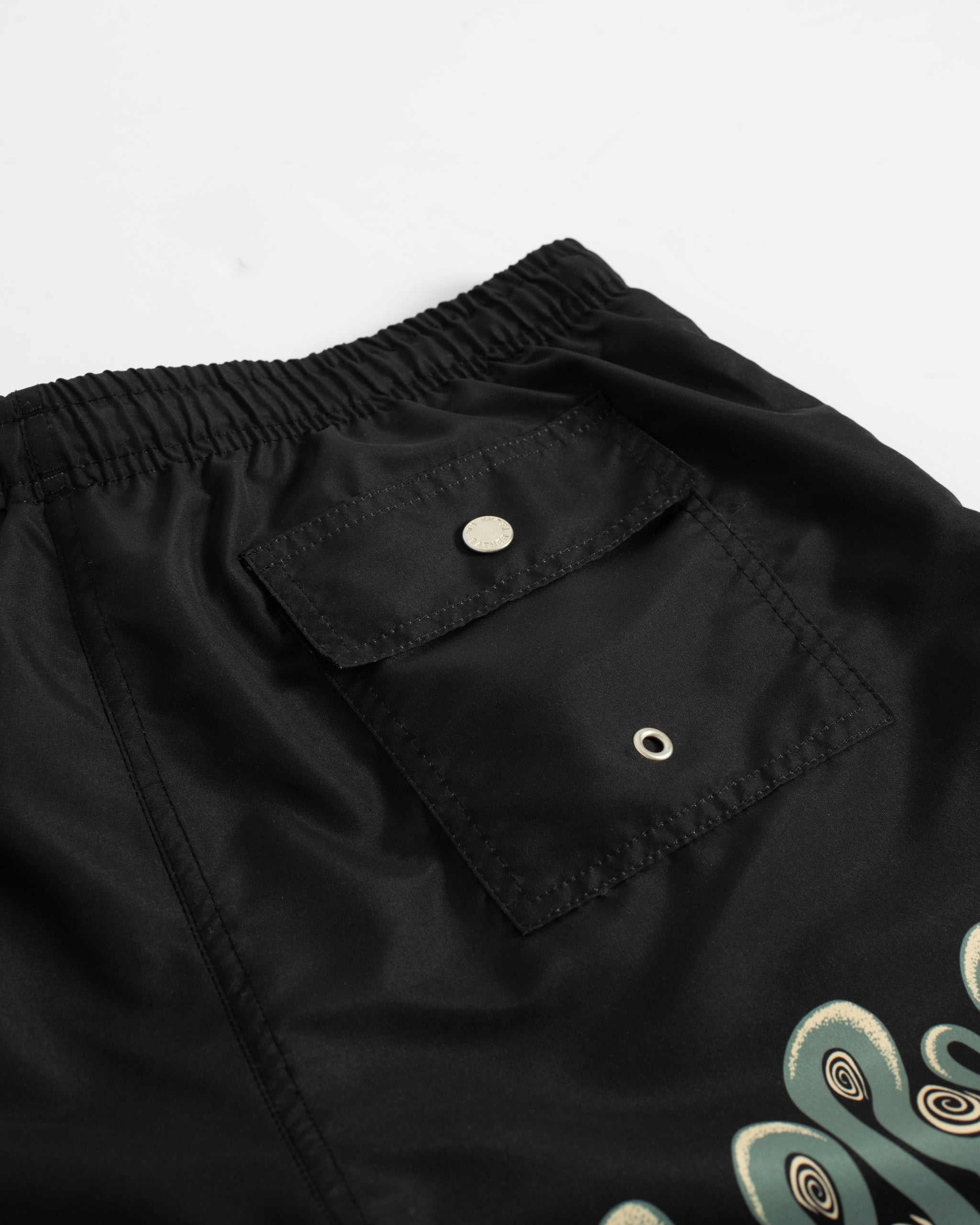 Close-up of men’s black swim trunks featuring a graphic snake border print and back pocket with button closure