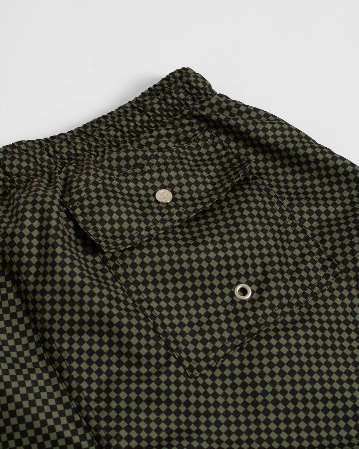 Close-up of a dark olive green and black checkerboard patterned garment, showing part of a collar, button closure, and fabric texture on a light gray background.