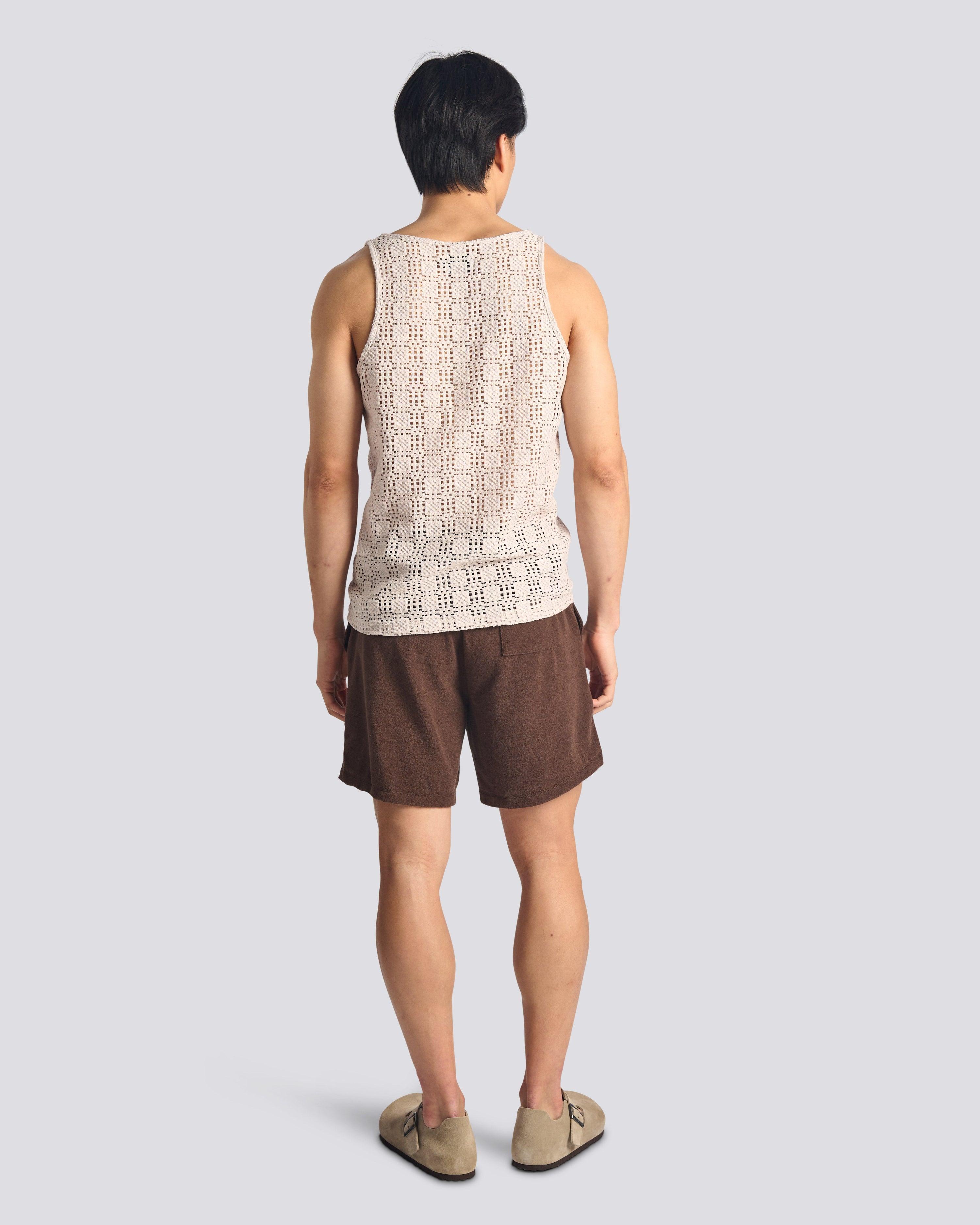 Back of Male model wearing the Bather Men's Lattice Tank Top in a relaxed fit, showcasing the breathable open-weave fabric for a summer resort look with brown shorts