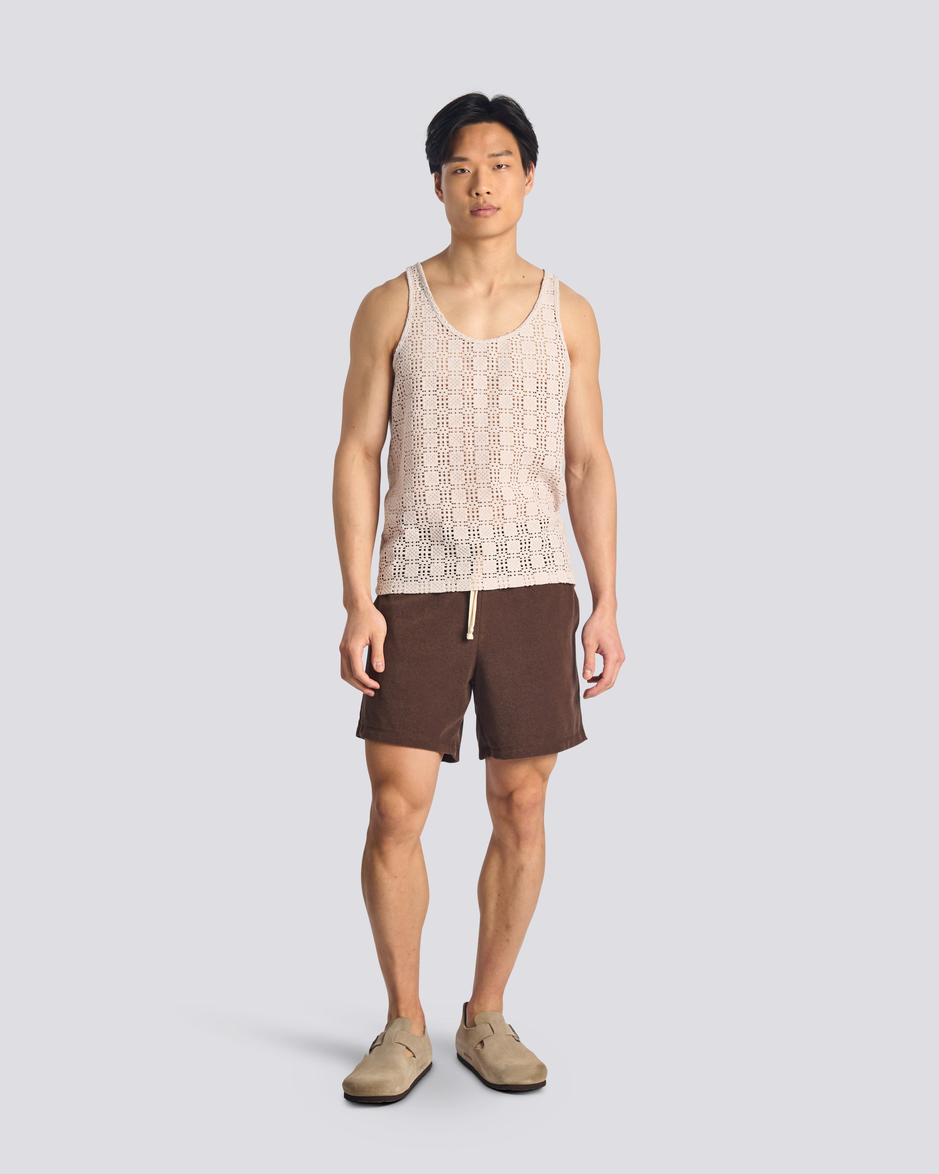 Male model wearing the Bather Men's Lattice Tank Top in a relaxed fit, showcasing the breathable open-weave fabric for a summer resort look with brown shorts.