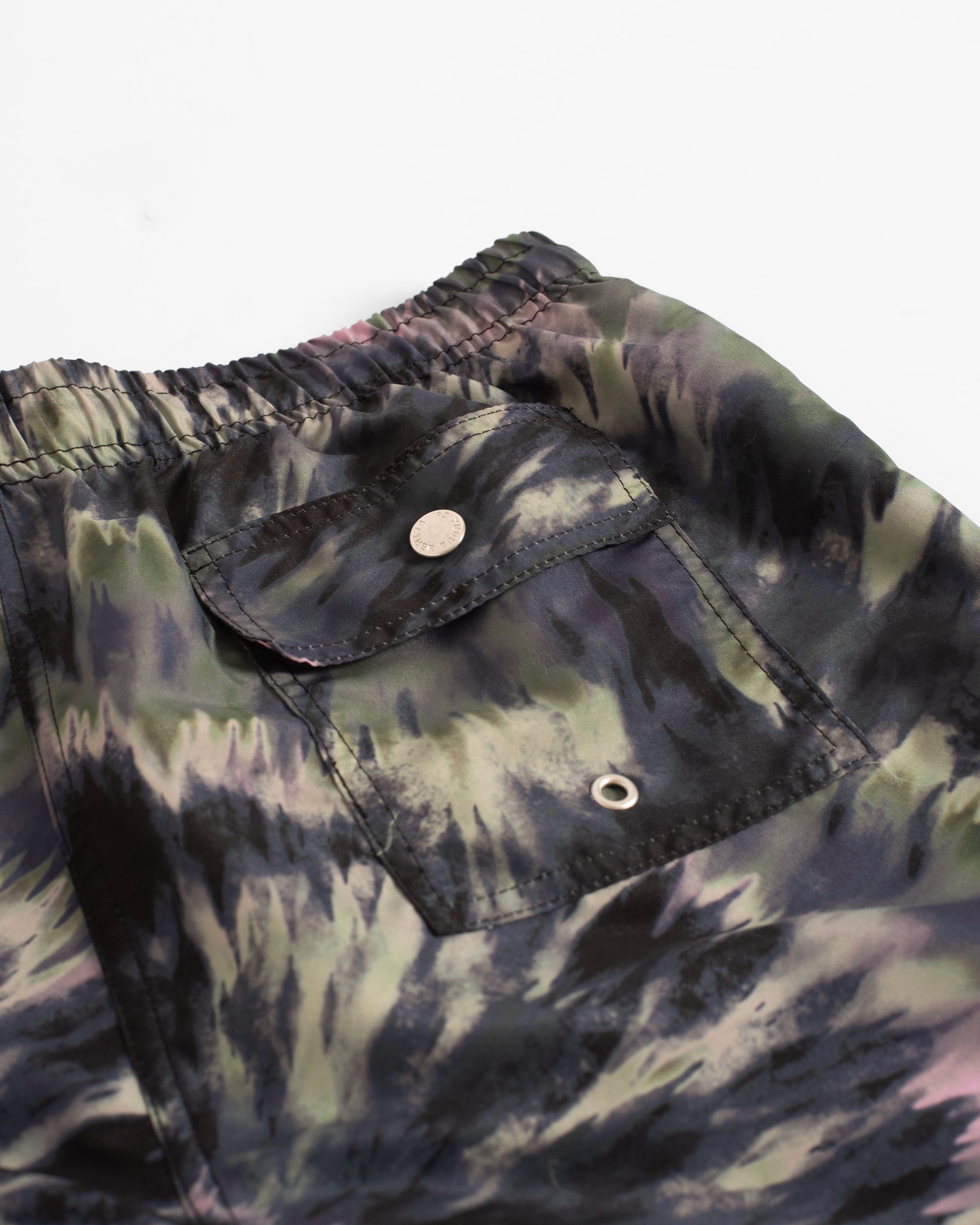 Close-up of men’s abstract tie-dye swim trunks in black, green, cream, and pink, with button closure back pocket
