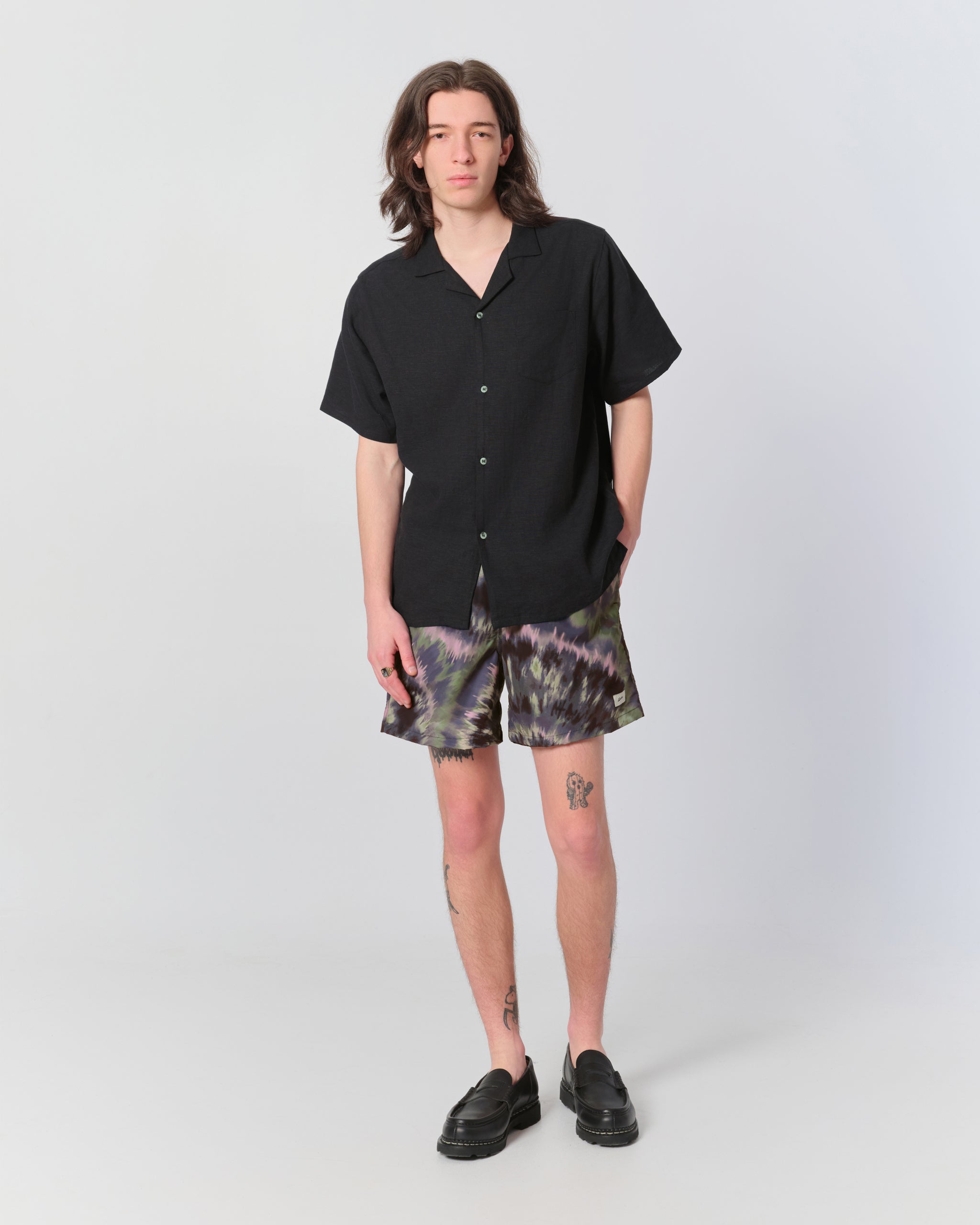 Man wearing abstract tie-dye swim trunks in dark green and plum tones and matching Bather linen camp shirt 