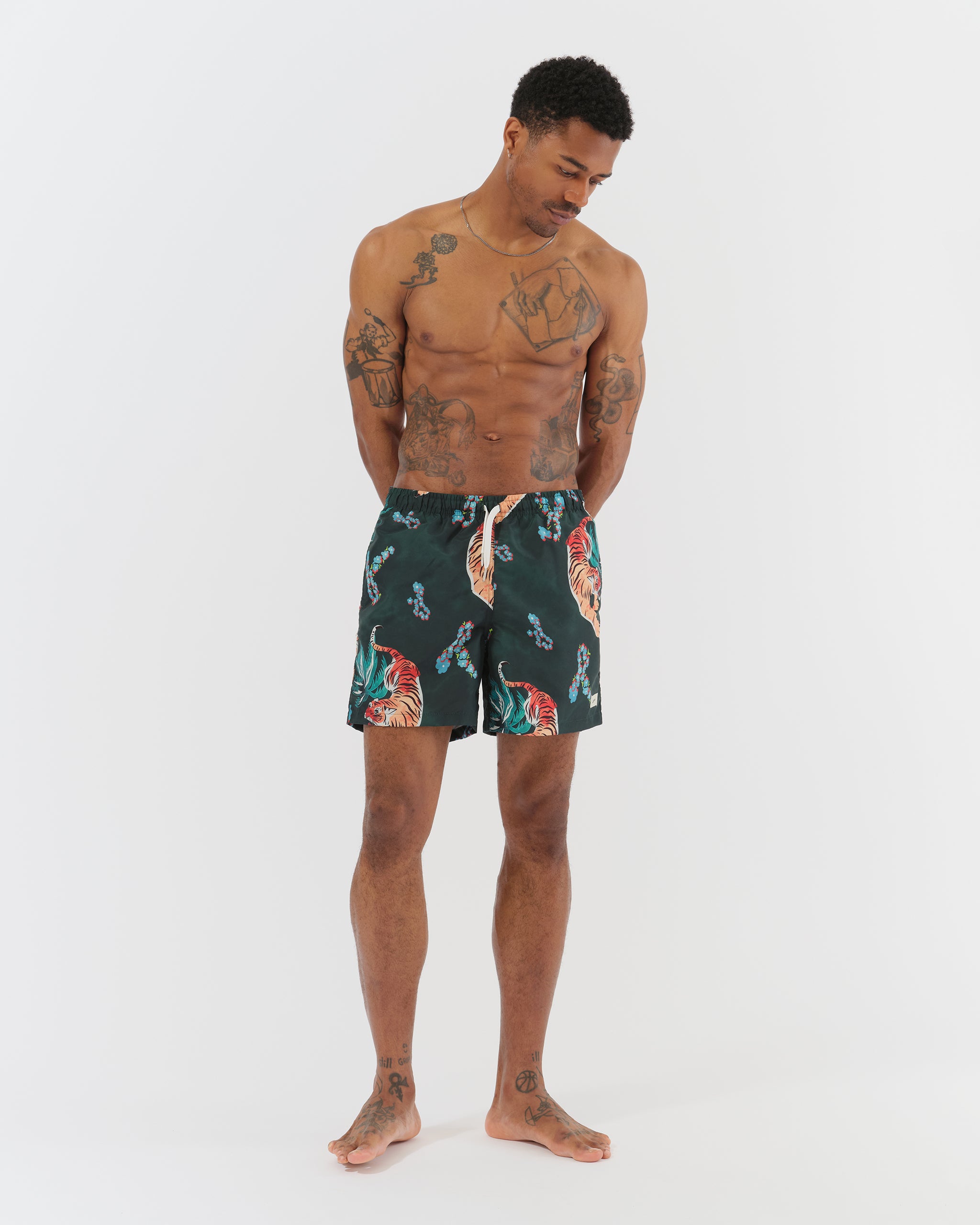 Shirtless tattooed man wearing all over print swim trunks with a forest and tigers in dark green and warm orange tones