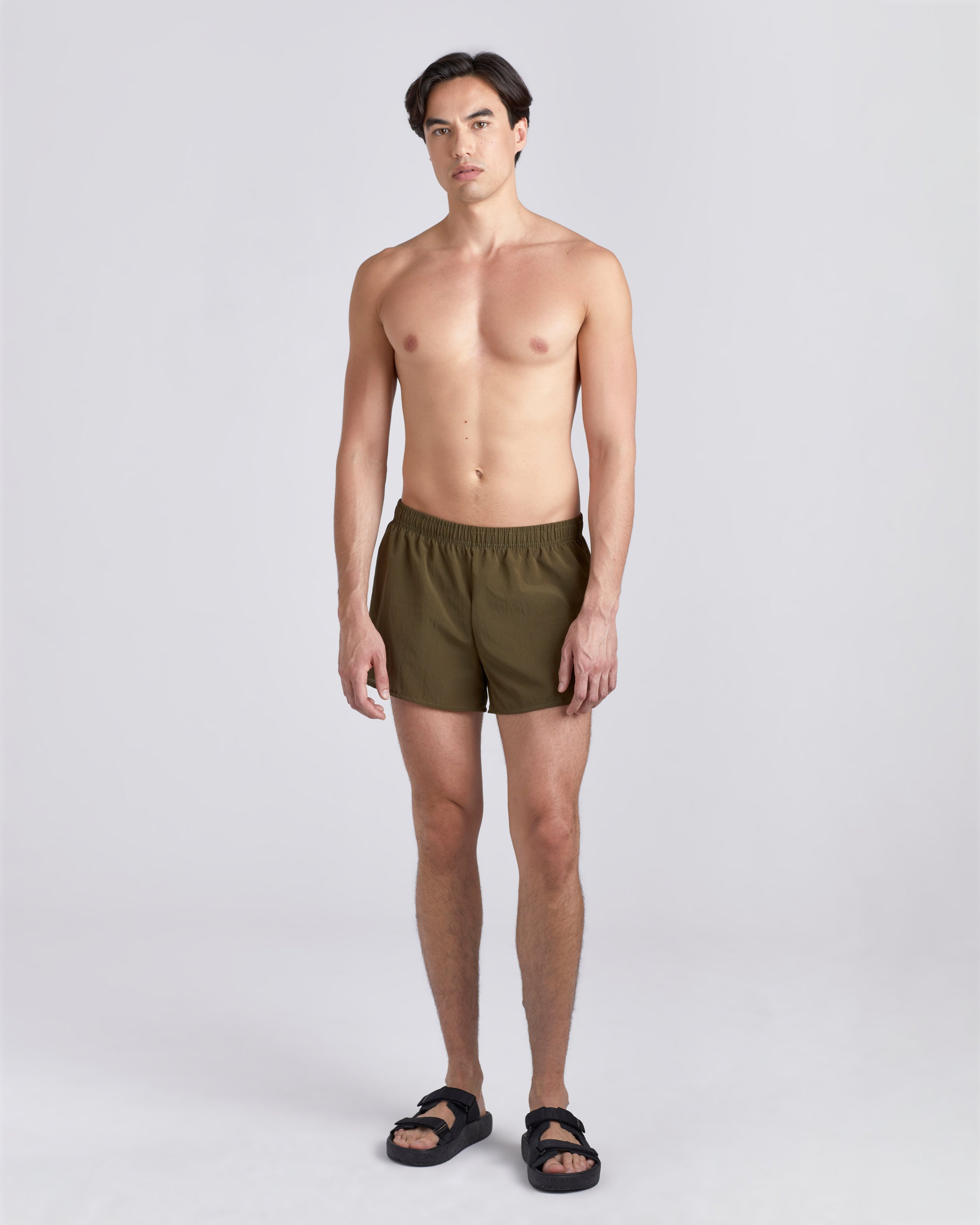Front view of shirtless model wearing sandals and 3.5 inch inseam Bather Active Trunk in green featuring lightweight recycled Japanese DotAir nylon with stretch fabric and compression liner