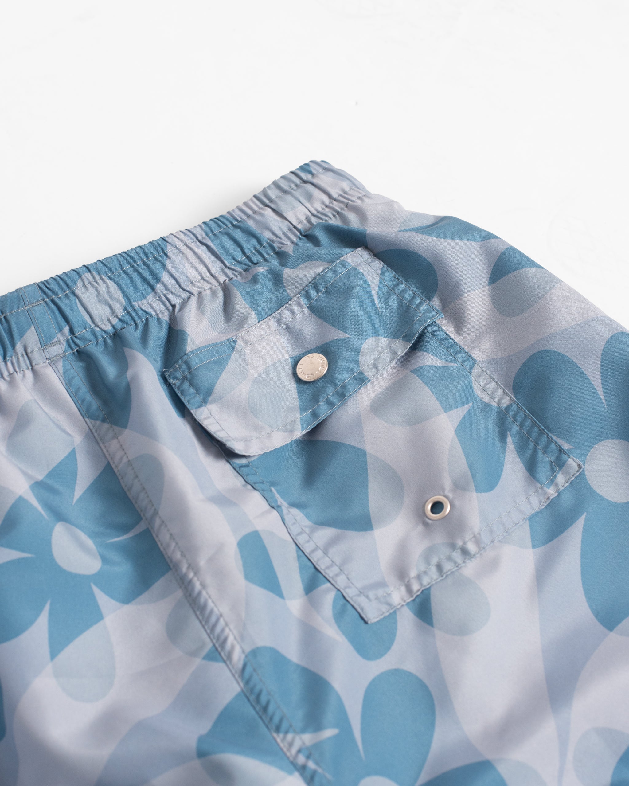 Close-up of men’s all over print swim trunks with layered floral pattern in light blue and dark blue tones featuring button closure back pocket