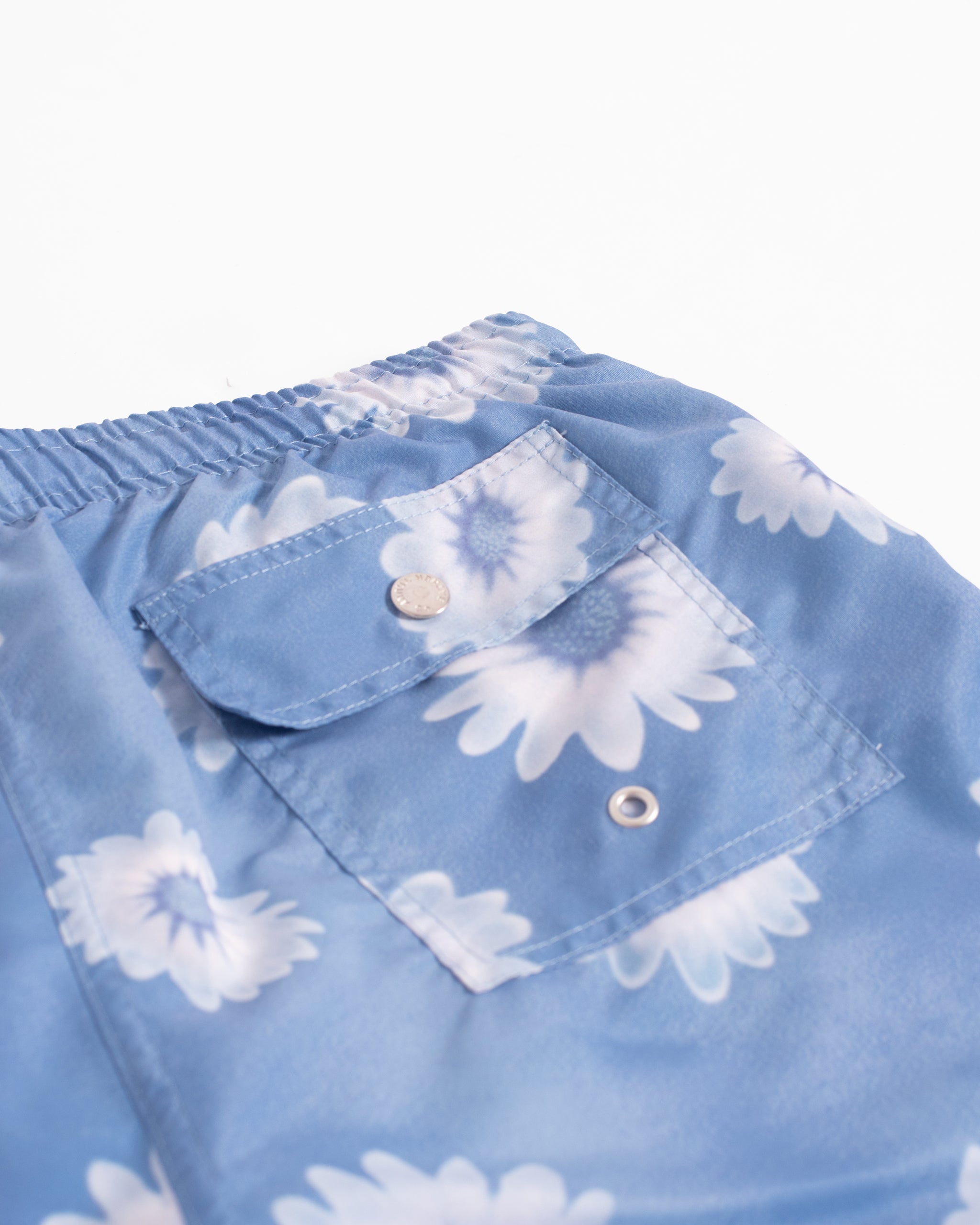 Detail view of men’s daisy pattern all over print swim trunks in light blue featuring button closure back pocket