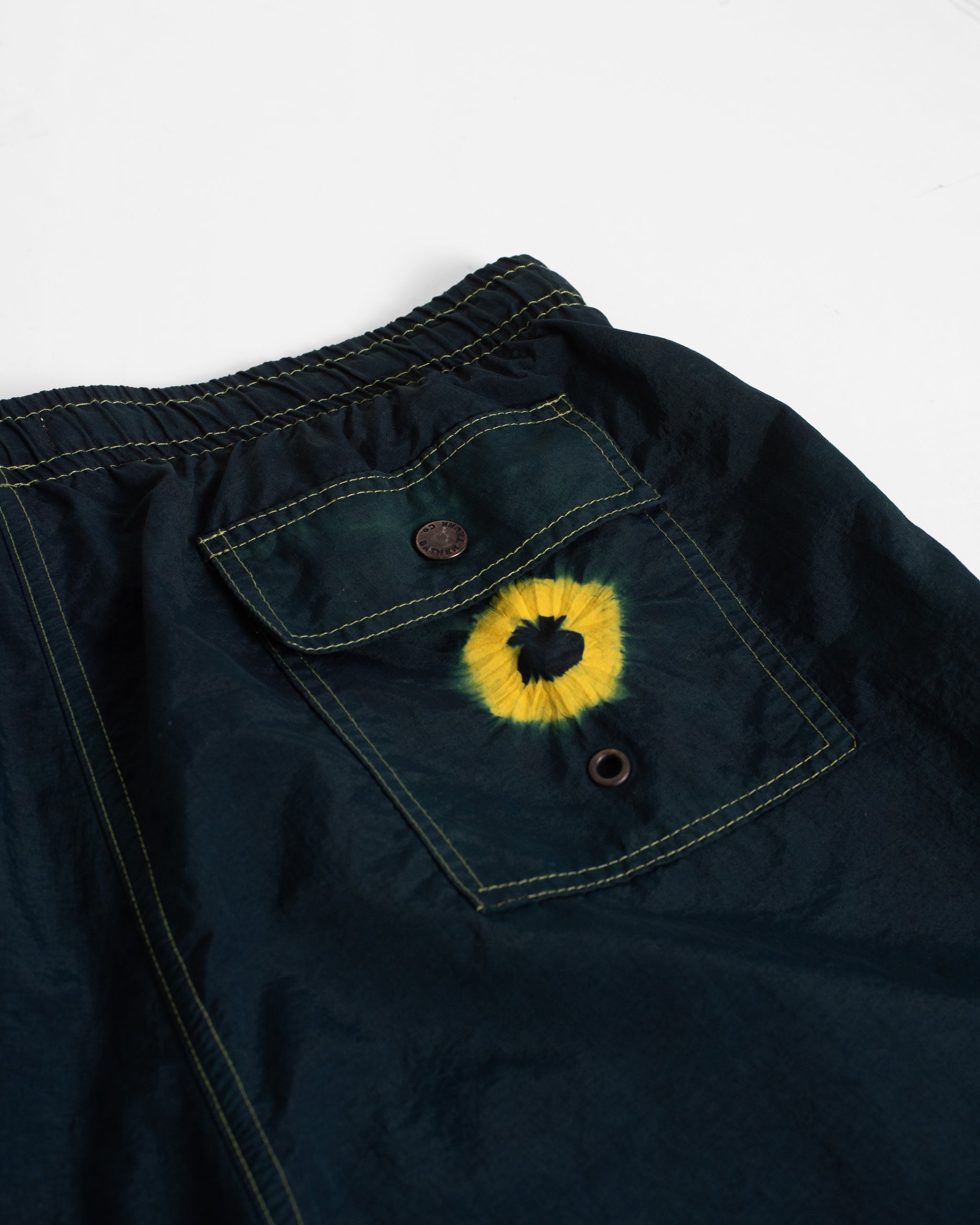 Navy blue swim trunks with a yellow tie dye emblem on a white background
