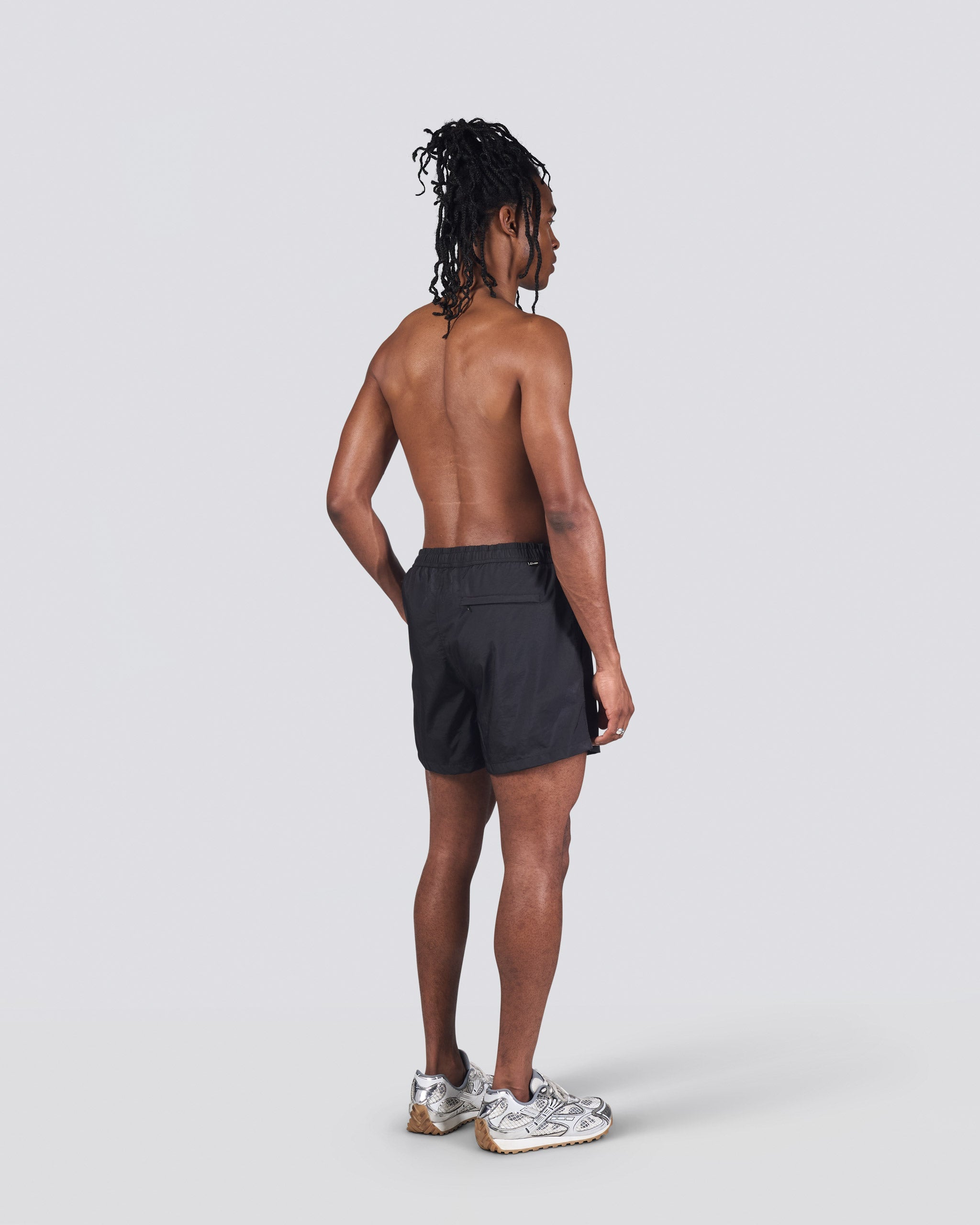 Back of shirtless man wearing shiny black swim trunk featuring an elastic drawstring waistband on white background