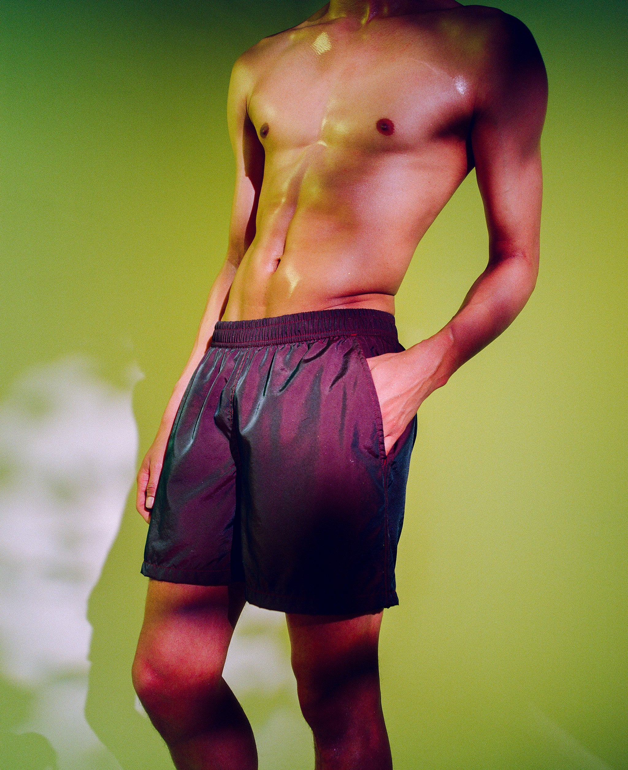 Torso and legs of shadowy shirtless man posing in shiny plum swim trunk featuring an elastic drawstring waistband on red lit background