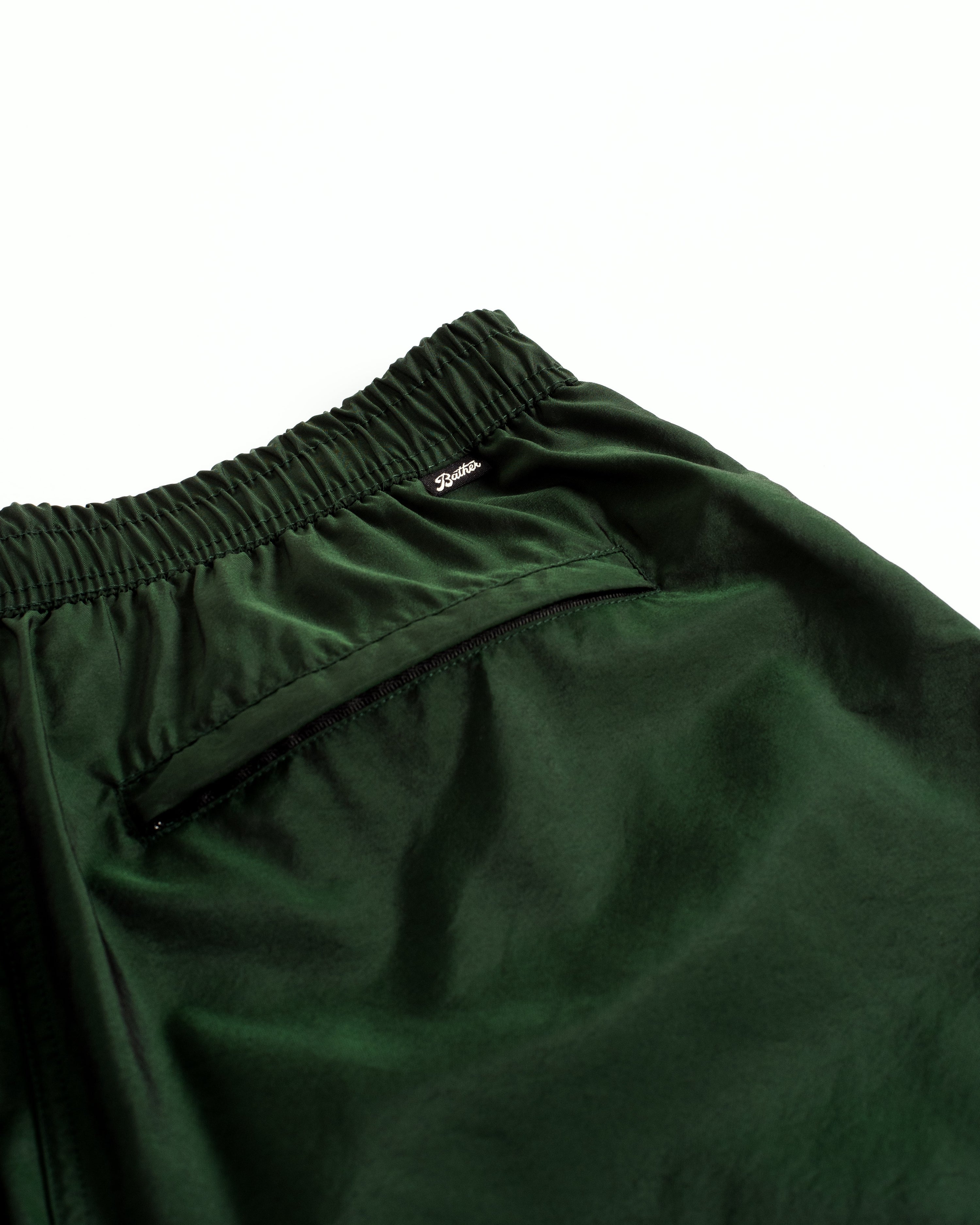 Detail close up view of men's shiny swim trunk in dark green featuring zipper closure back pocket on white background