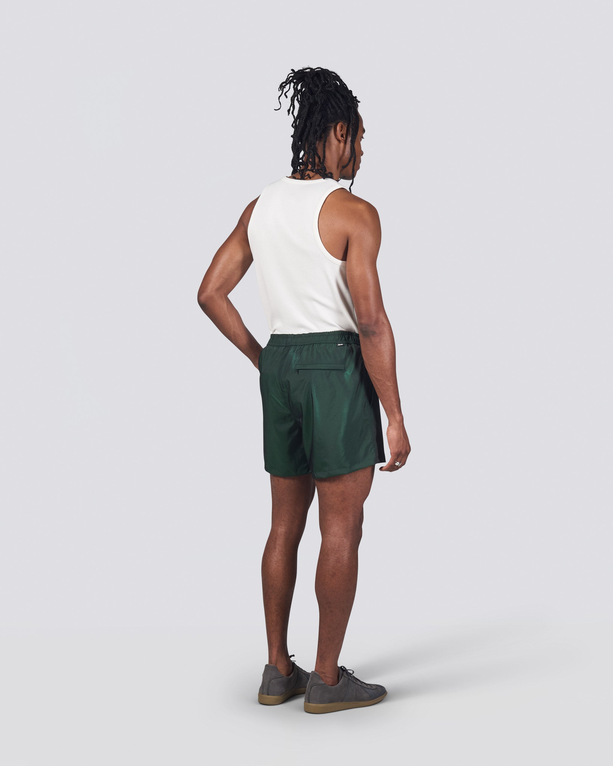 Back of man in white tank top wearing shiny dark green swim trunk featuring an elastic drawstring waistband on white background