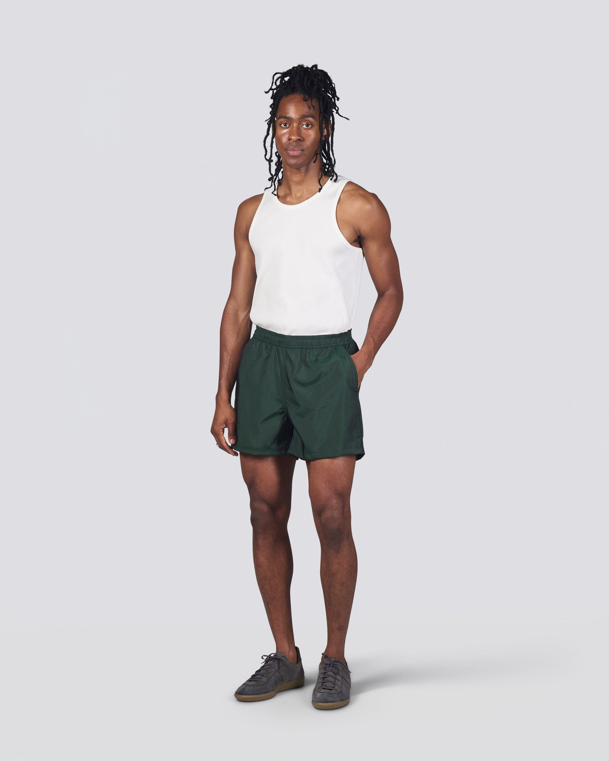 Man in white tank top wearing shiny dark green swim trunk featuring an elastic drawstring waistband on white background