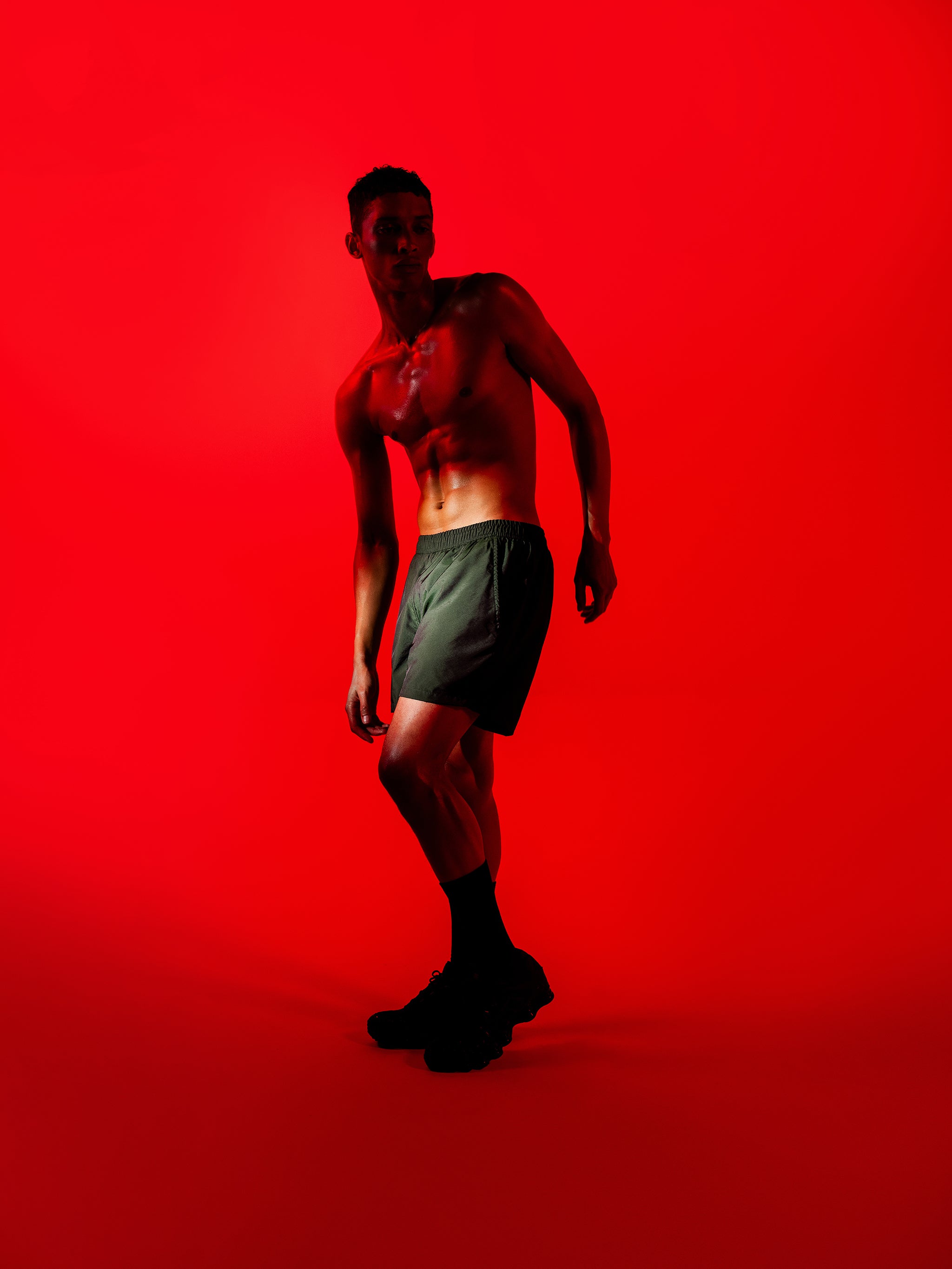 Shadowy shirtless man posing in shiny dark green swim trunk featuring an elastic drawstring waistband on red lit background.
