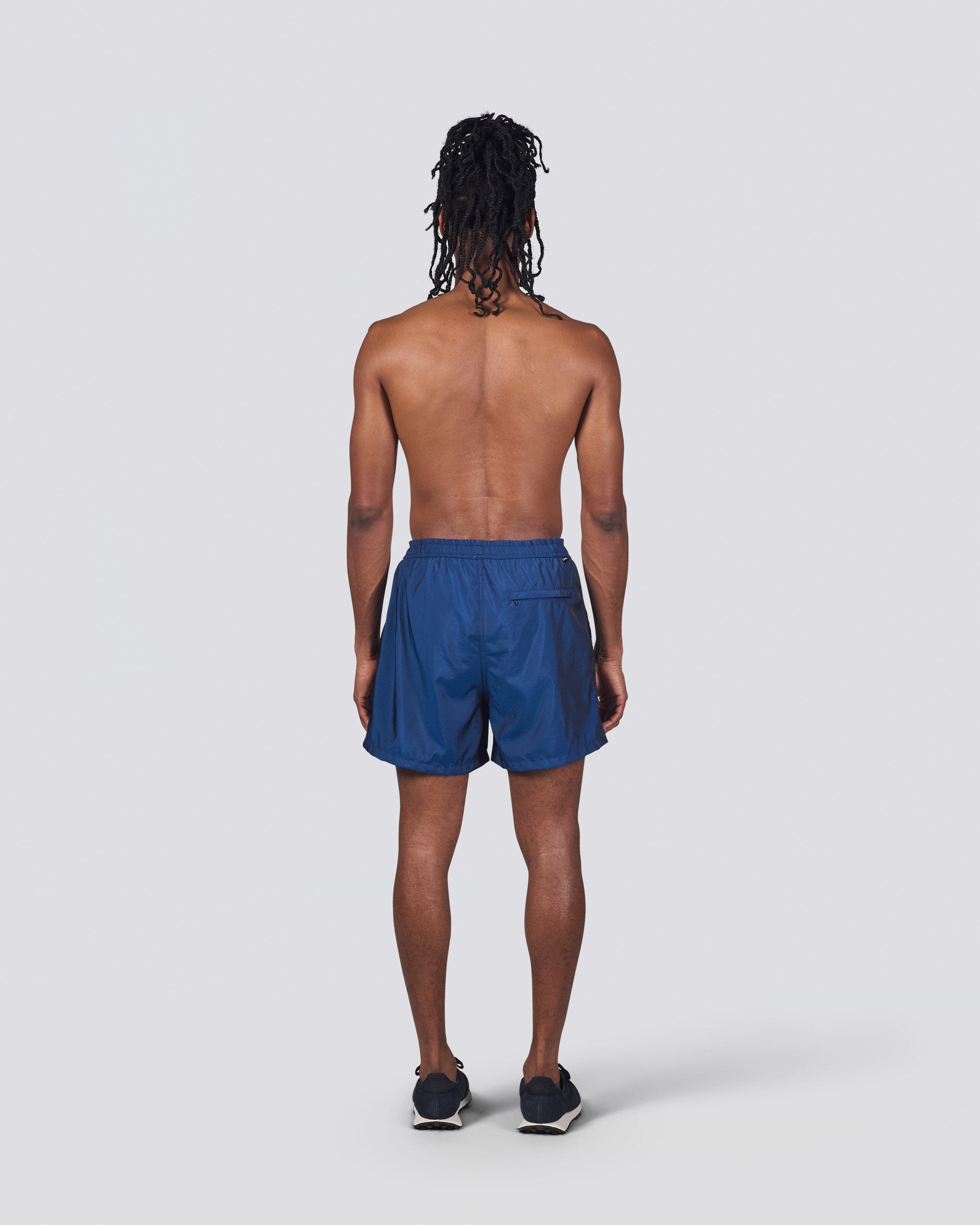 Back of shirtless man wearing shiny dark blue swim trunk featuring an elastic drawstring waistband on white background