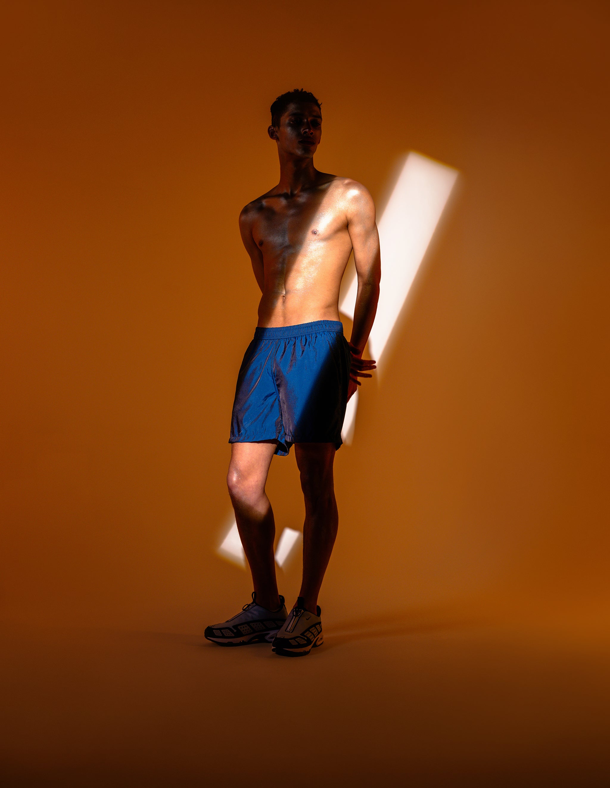 Shadowy shirtless man posing in shiny dark blue swim trunk featuring an elastic drawstring waistband on orange lit background.