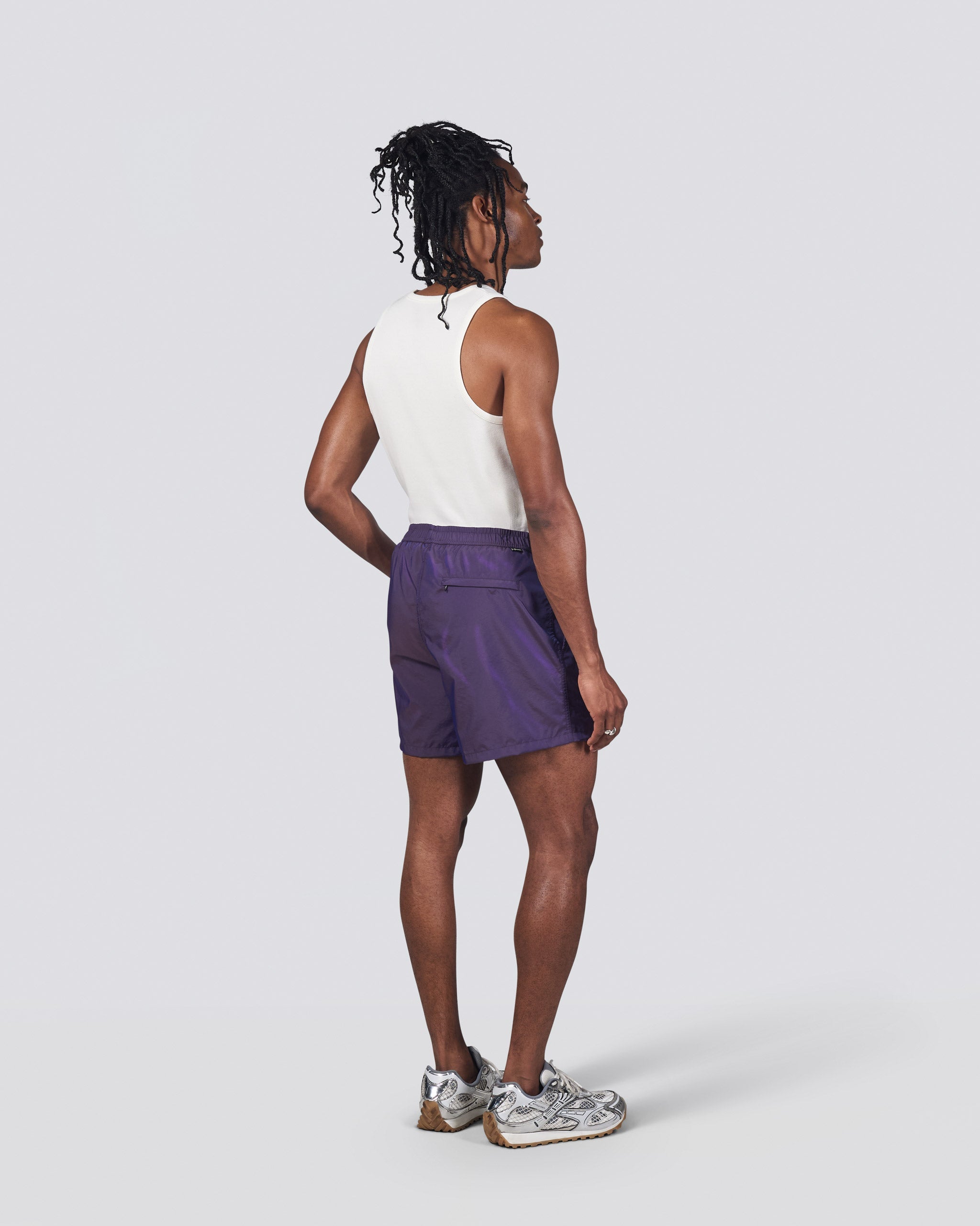 Back of man in white tank top wearing shiny dark purple swim trunk featuring an elastic drawstring waistband on white background