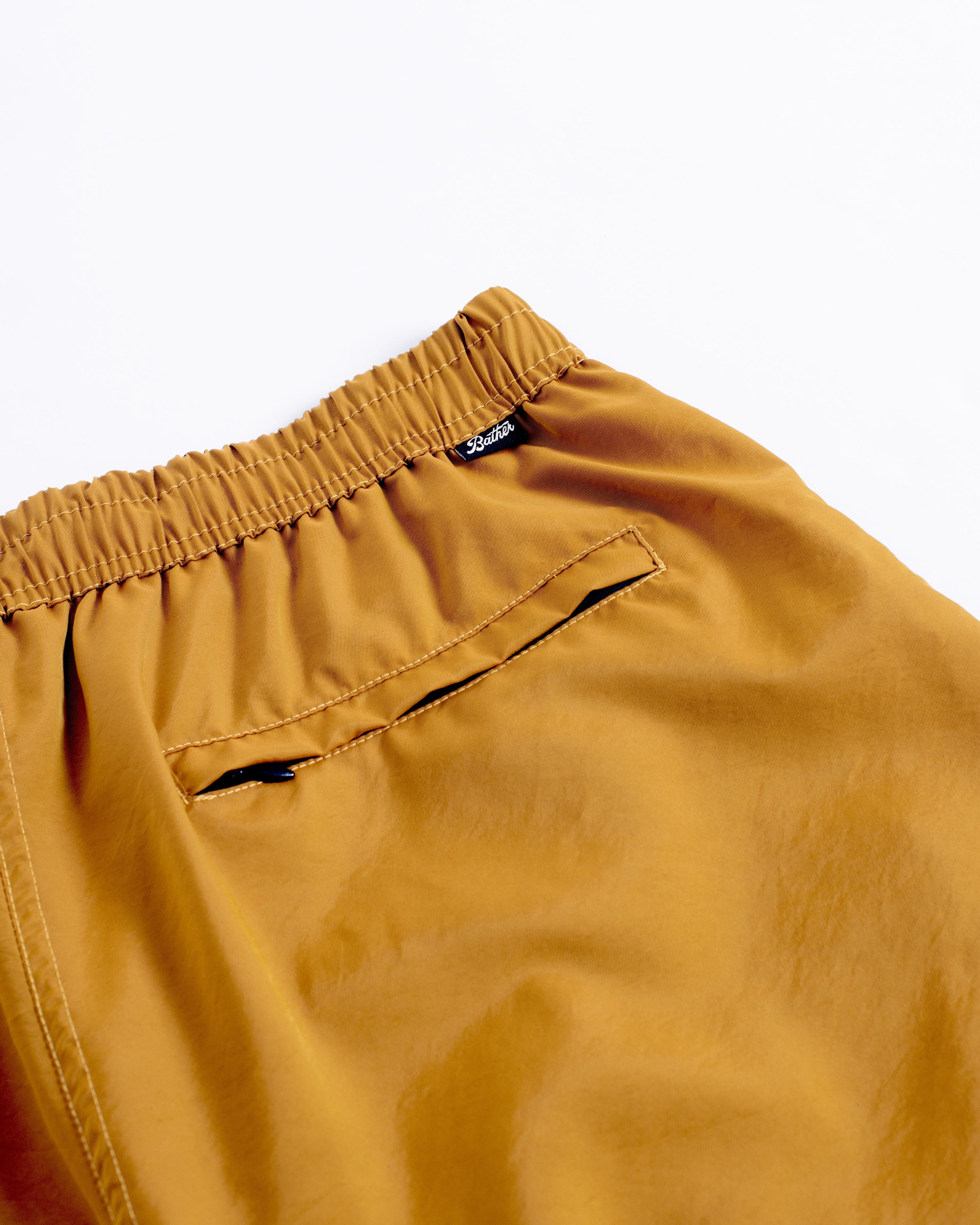 Detail close up view of men's shiny swim trunk in mustard yellow featuring zipper closure back pocket on white background