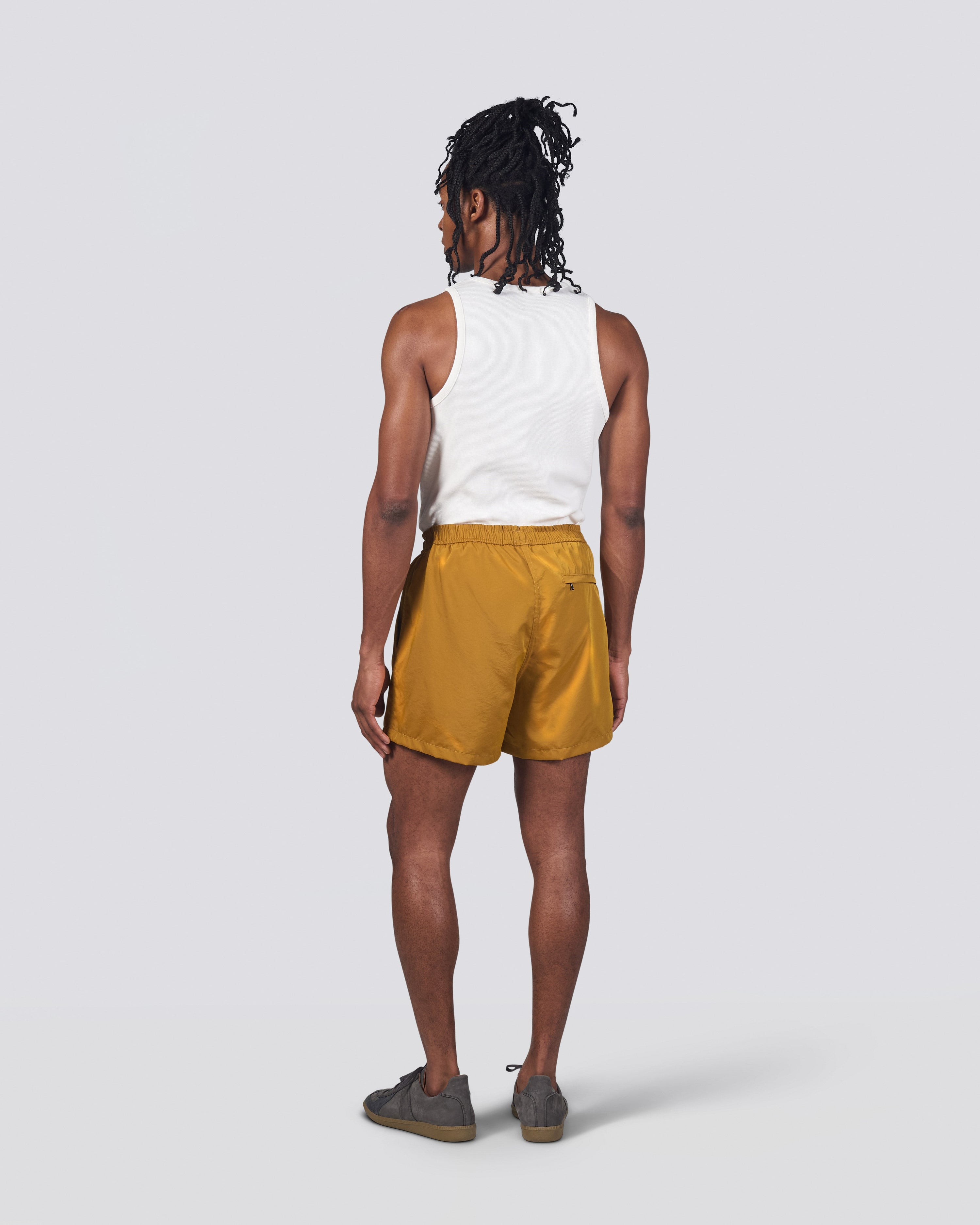 Back of man in white tank top wearing shiny mustard yellow swim trunk featuring an elastic drawstring waistband on white background