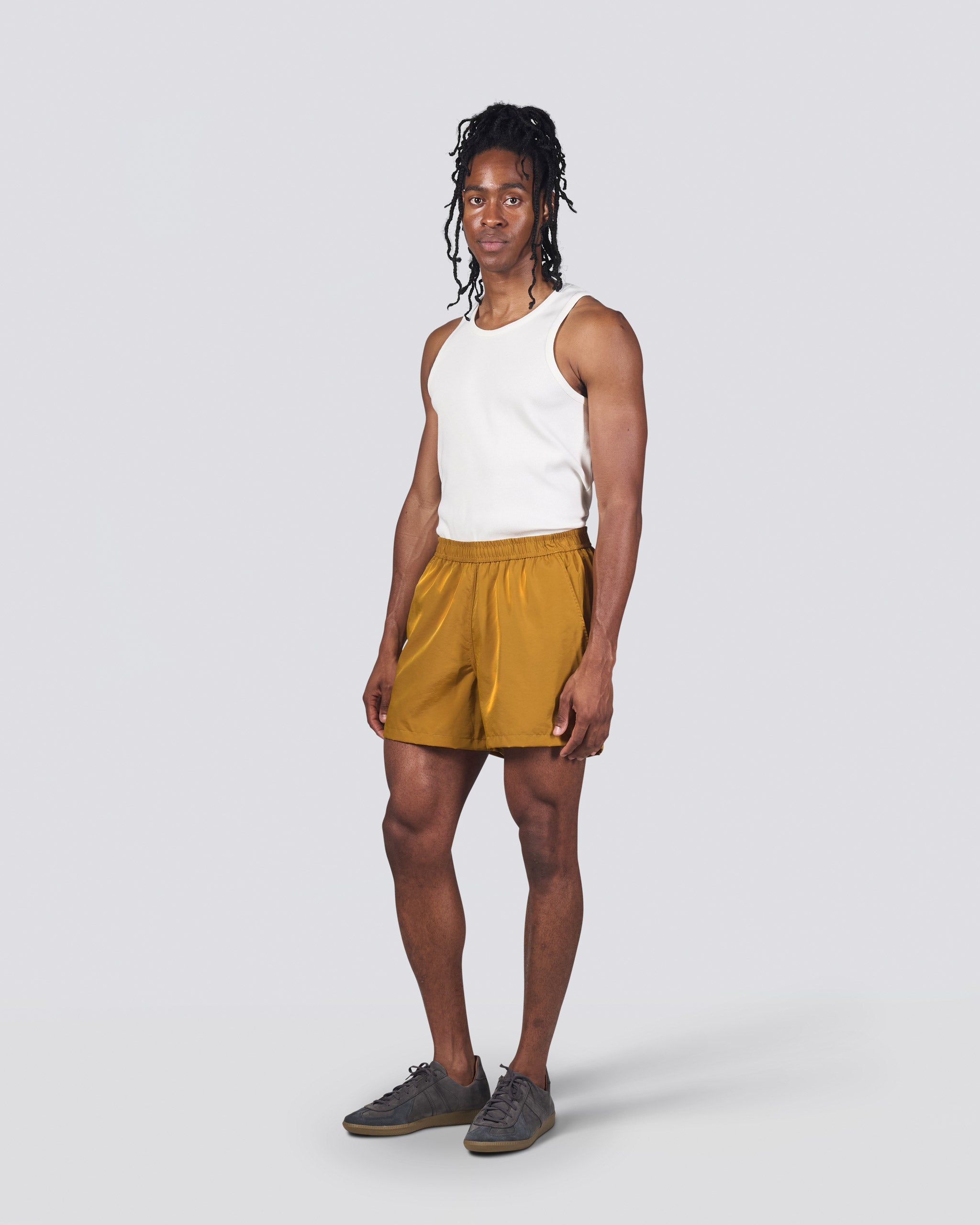 Man in white tank top wearing shiny mustard yellow swim trunk featuring an elastic drawstring waistband on white background
