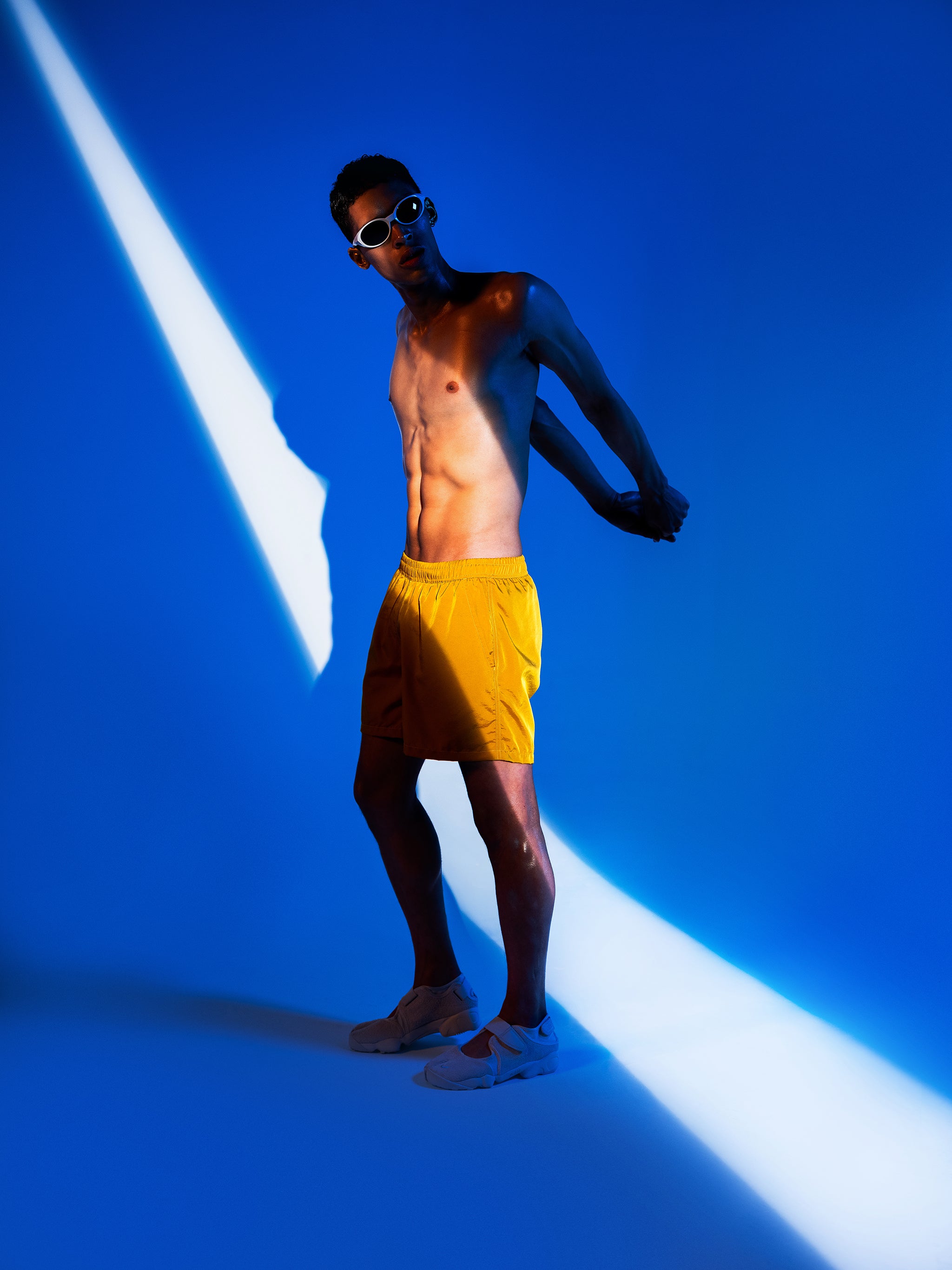 Shadowy shirtless man posing in shiny mustard yellow swim trunk featuring an elastic drawstring waistband on blue lit background