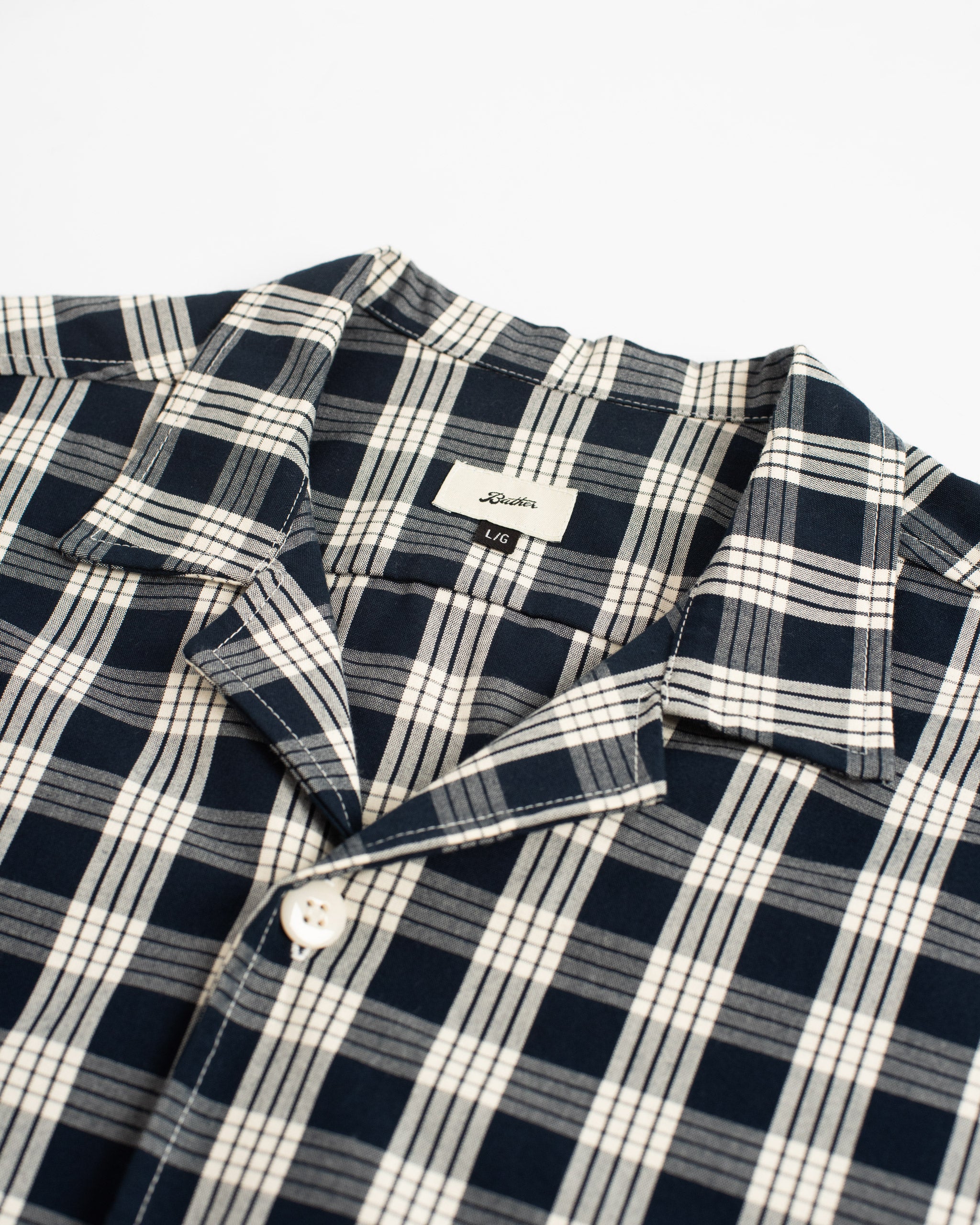 A close-up detail of the Bather Men's Admiral Palaka Traveler Camp Shirt, showing the navy and white Polynesian plaid fabric, the classic camp collar construction, and a white button.