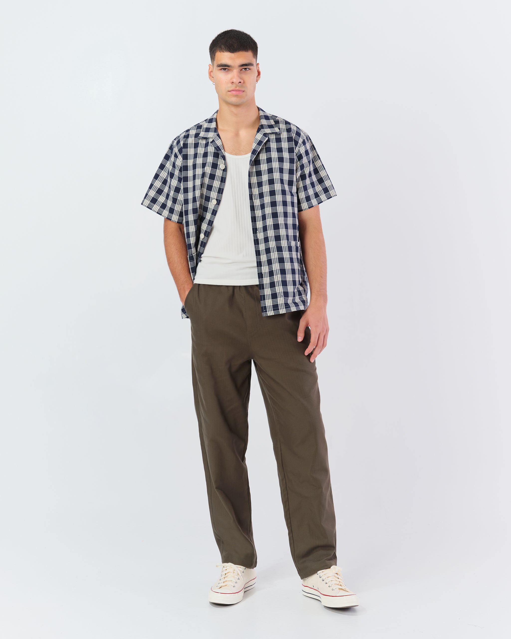 A model wearing the Bather Men's Admiral Palaka Traveler Camp Shirt, styled open over a white tank top and olive green trousers. The image highlights the navy and white Polynesian plaid pattern and the shirt's relaxed, short-sleeve silhouette.