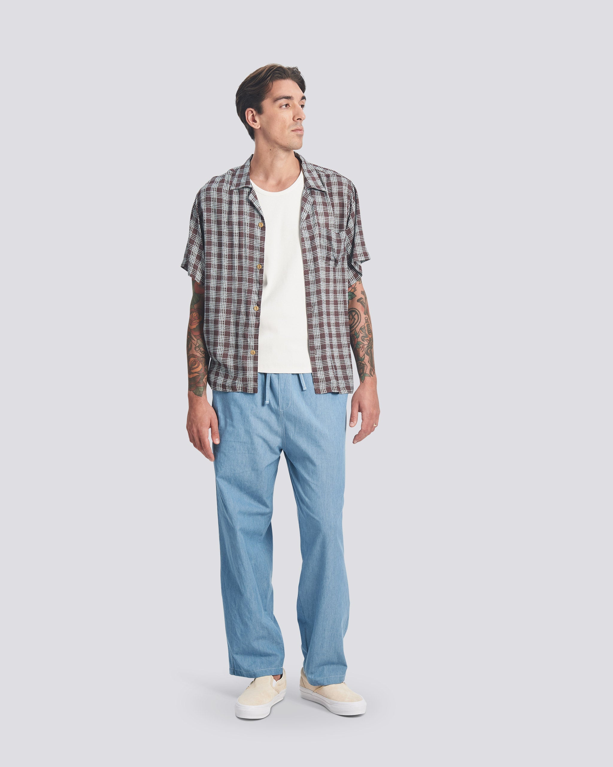Model wearing the Bather Men's Amethyst Tartan Camp Shirt styled open over a white t-shirt and light blue trousers. The shot shows the relaxed, boxy fit and the purple and blue hand-drawn plaid pattern.