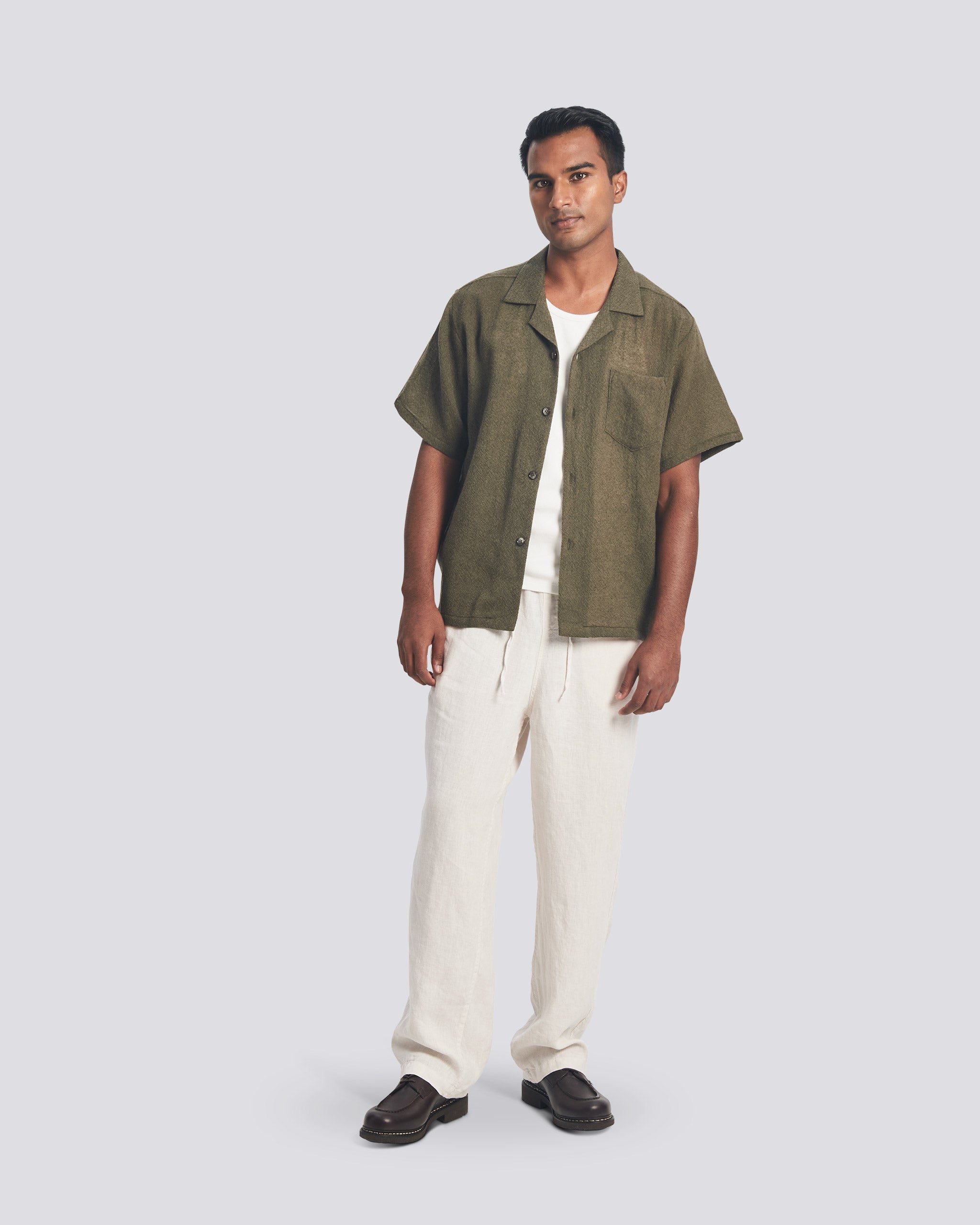 Full-length shot of a model wearing the Bather Men's Army Wool Gauze Camp Shirt styled open over a white t-shirt and cream-colored trousers. The image showcases the dark olive Japanese wool texture and the shirt's relaxed, boxy fit.