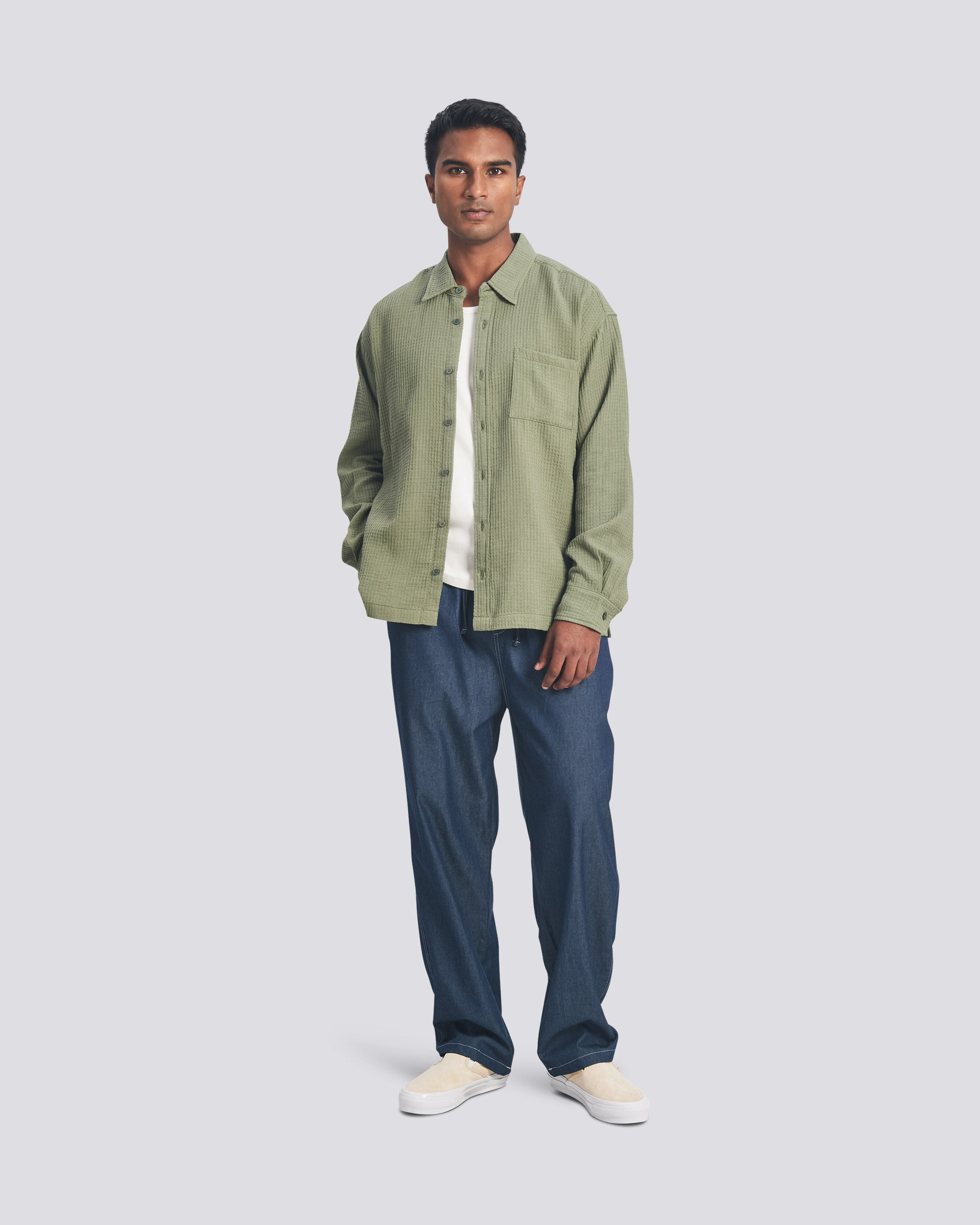 A model wearing the Bather Men's Artichoke Waffle Leisure Shirt, styled open over a white undershirt and dark navy trousers. The image showcases the oversized fit and the distinct textured waffle weave of the olive green cotton-linen fabric.
