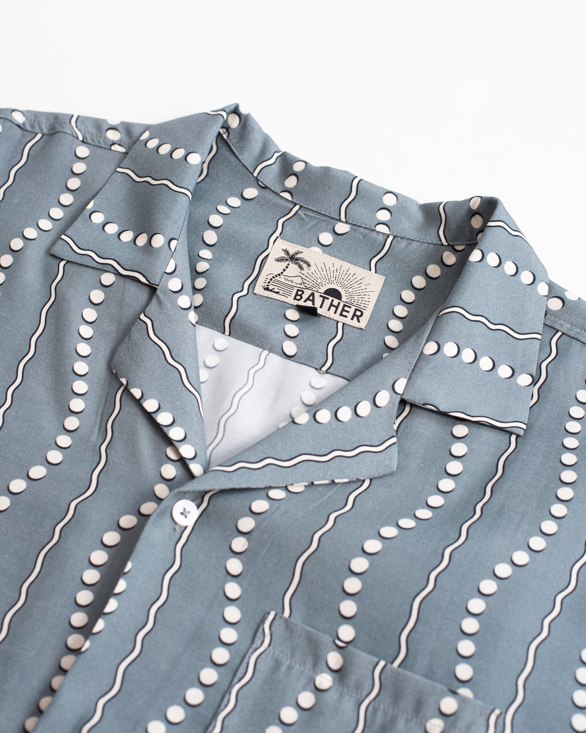 A close-up detail of the Bather Men's Ash Tidal Current Camp Shirt, showcasing the steel blue and white wavy vertical stripe motif, the classic camp collar, and the interior Bather brand label.