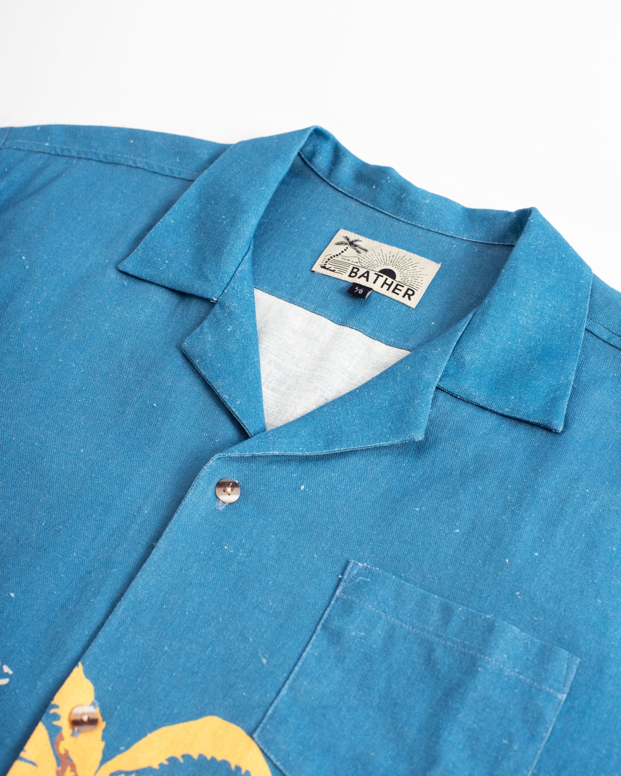 A close-up detail of the Bather Men's Baltic Trippin' Beach Camp Shirt, highlighting the textured blue linen fabric and the retro Polynesian-inspired beach scene print featuring palm trees and tropical motifs.