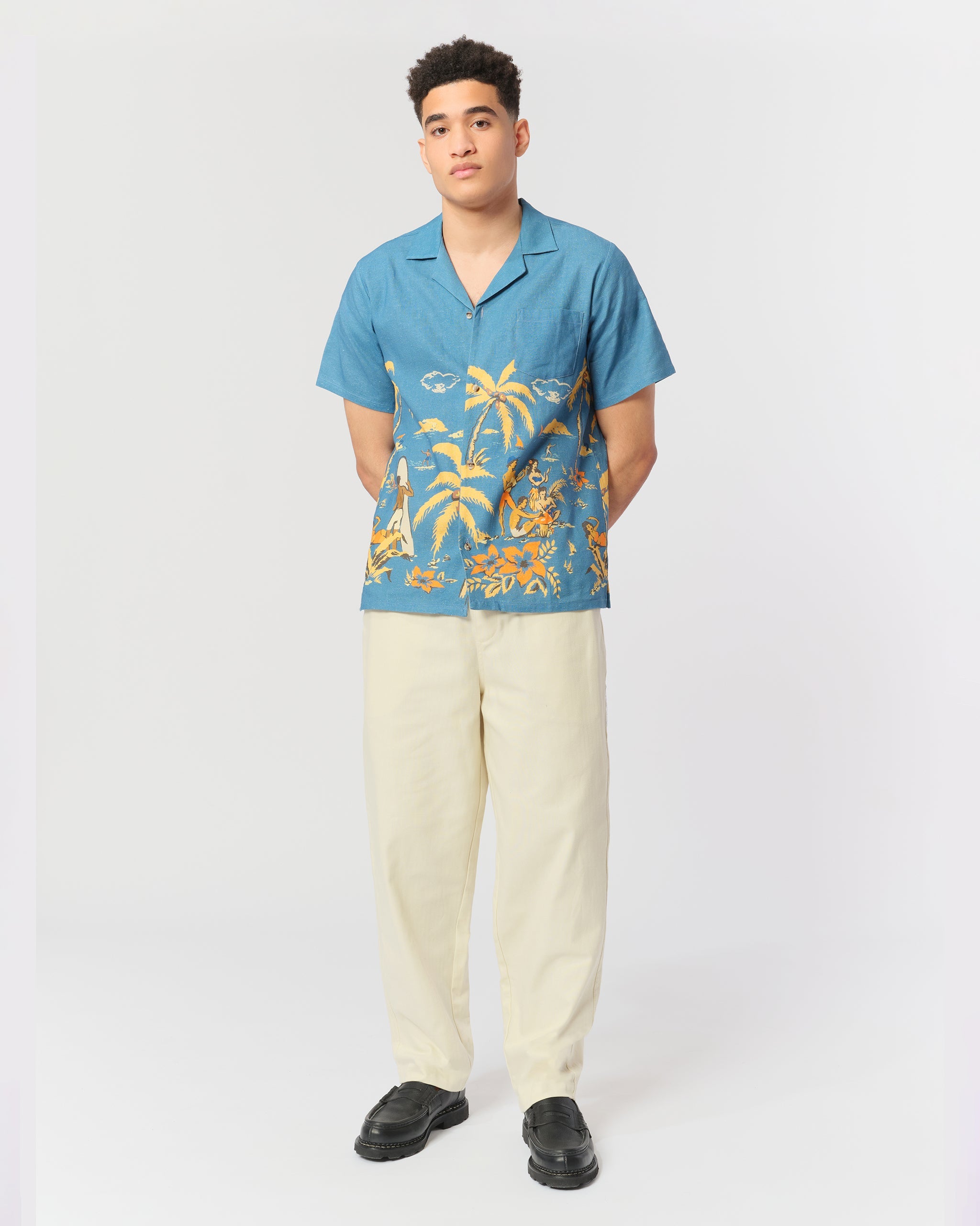 A model wearing the Bather Men's Baltic Trippin' Beach Camp Shirt, styled with cream trousers and black loafers. The image showcases the relaxed fit and the blue linen fabric featuring a retro Polynesian-inspired beach scene print.