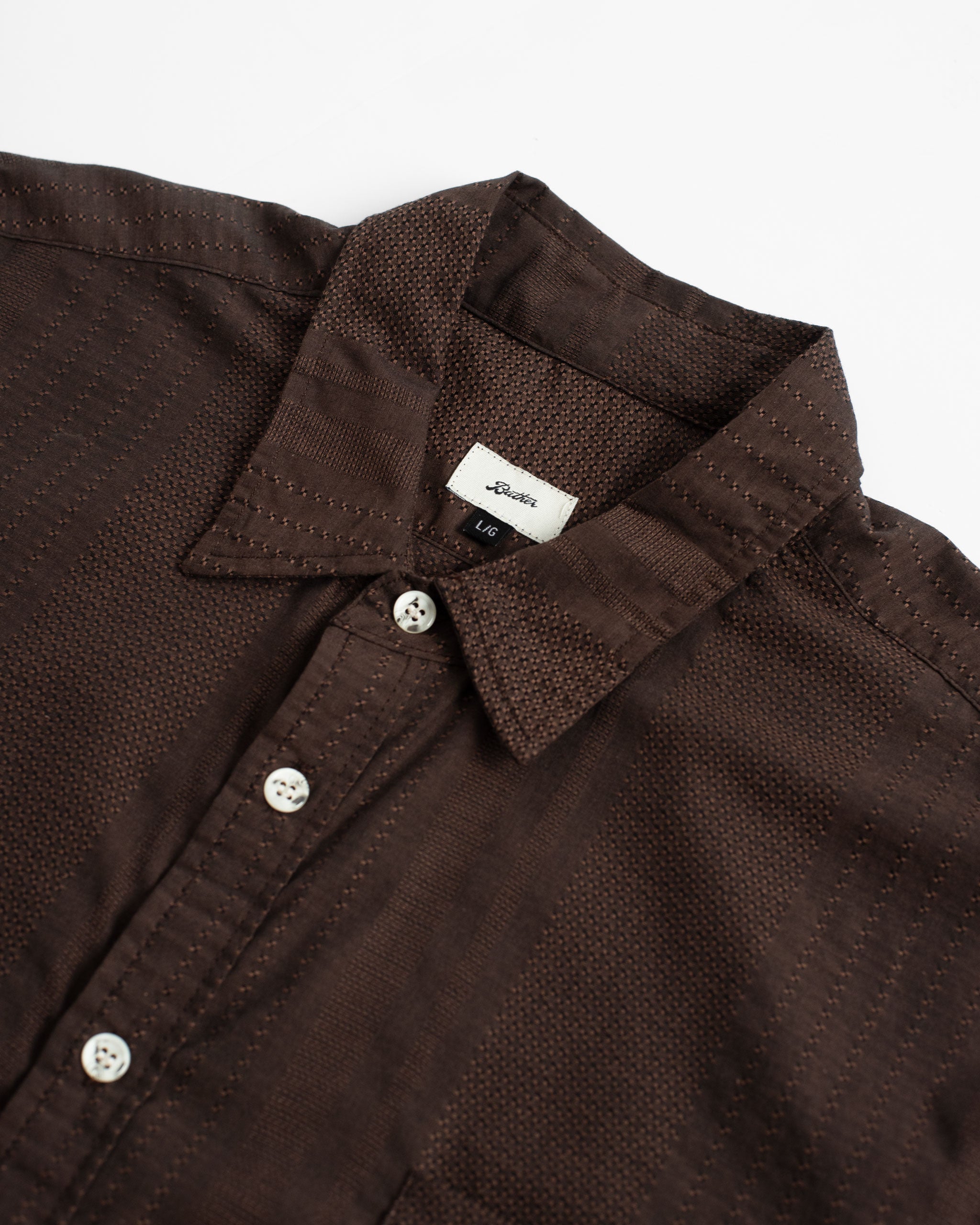 A close-up detail of the Bather Men's Bark Geo Dobby Leisure Shirt, showcasing the intricate checkered geometric embroidery on the dark brown Japanese cotton dobby fabric, the classic collar, and tonal white buttons.
