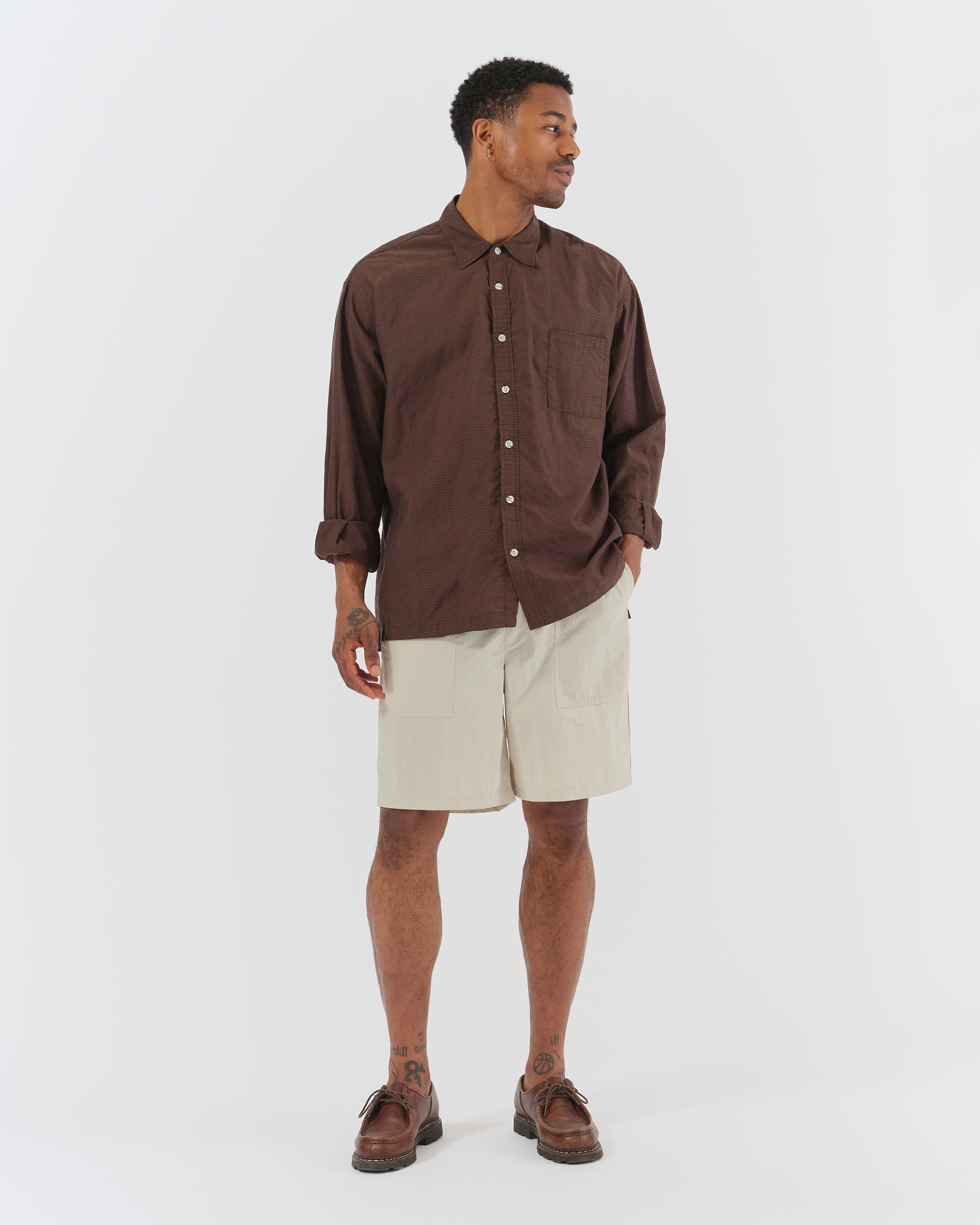 A model wearing the Bather Men's Bark Geo Dobby Leisure Shirt, styled with off-white shorts and brown leather loafers. The image showcases the shirt's oversized fit, long sleeves, and the intricate dark brown checkered geometric embroidery.