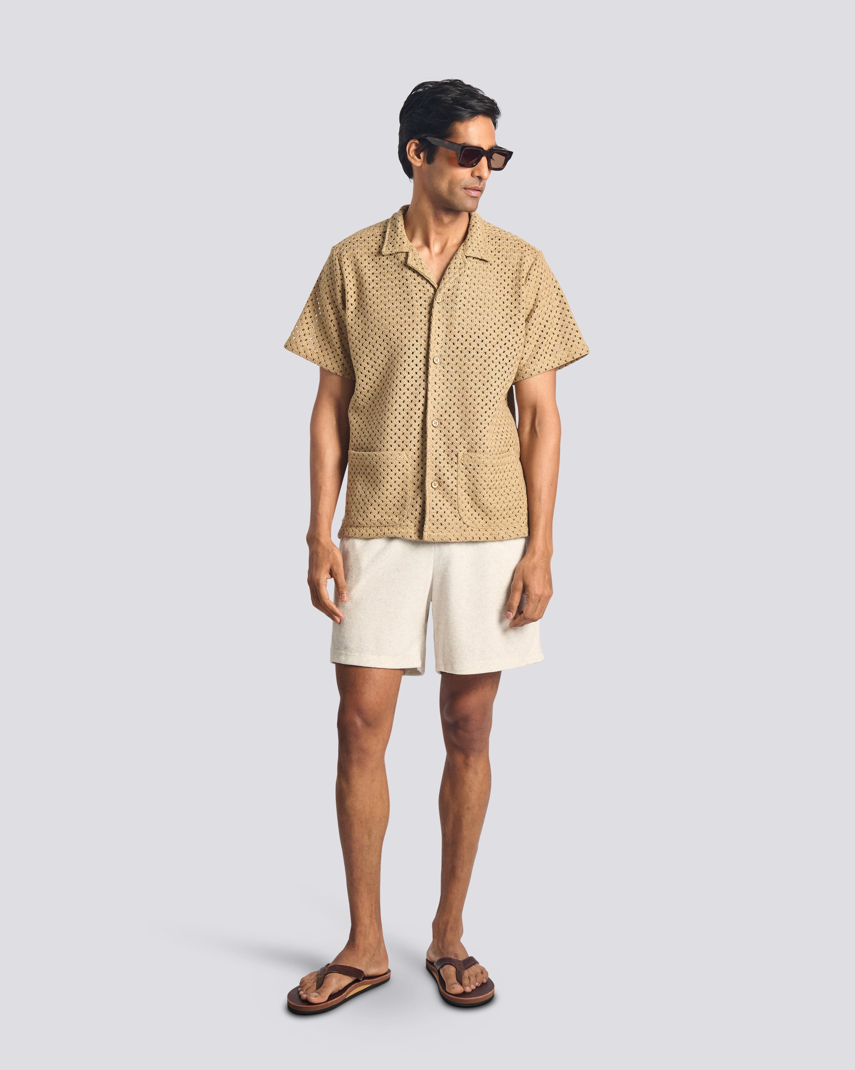 A model wearing the Bather Men's Beach Mesh Traveler Shirt, styled with white shorts and brown leather sandals. The image highlights the natural-colored Japanese cotton crocheted mesh fabric, the front patch pockets, and the shirt's relaxed, airy fit.