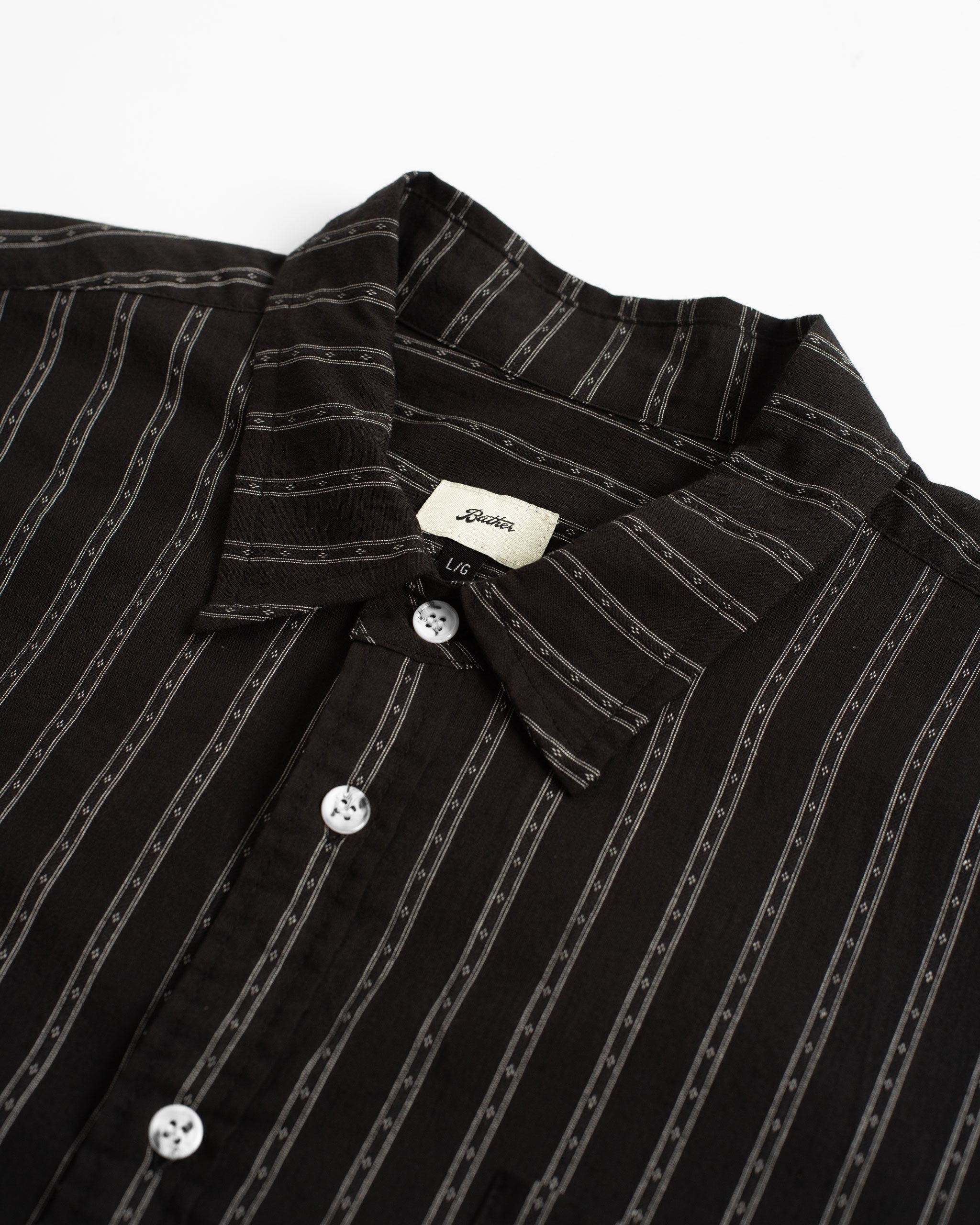 A close-up detail of the Bather Men's Black Dobby Leisure Shirt, highlighting the lightweight black Japanese cotton dobby fabric with a fine vertical stripe pattern, the classic collar, and white buttons.