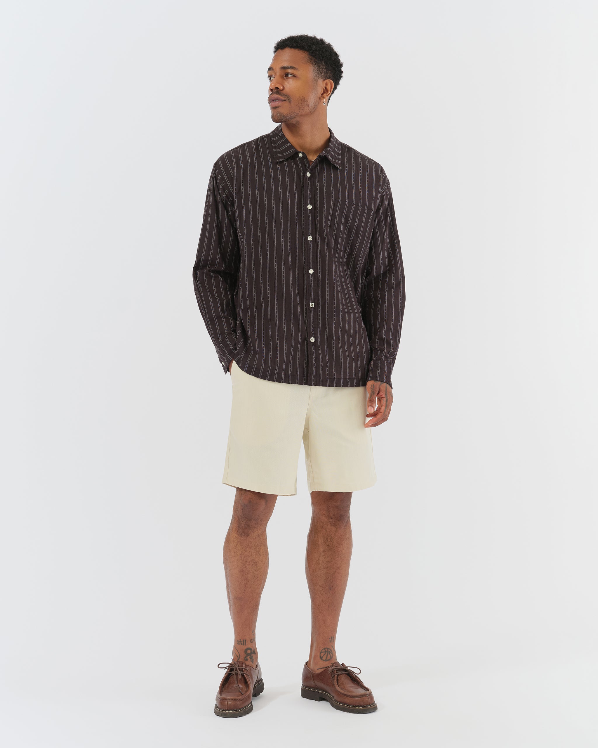 A model wearing the Bather Men's Black Dobby Leisure Shirt, styled with off-white shorts and brown leather loafers. The image highlights the shirt's oversized fit, long-sleeve silhouette, and the lightweight black Japanese cotton dobby fabric with a fine vertical stripe pattern.