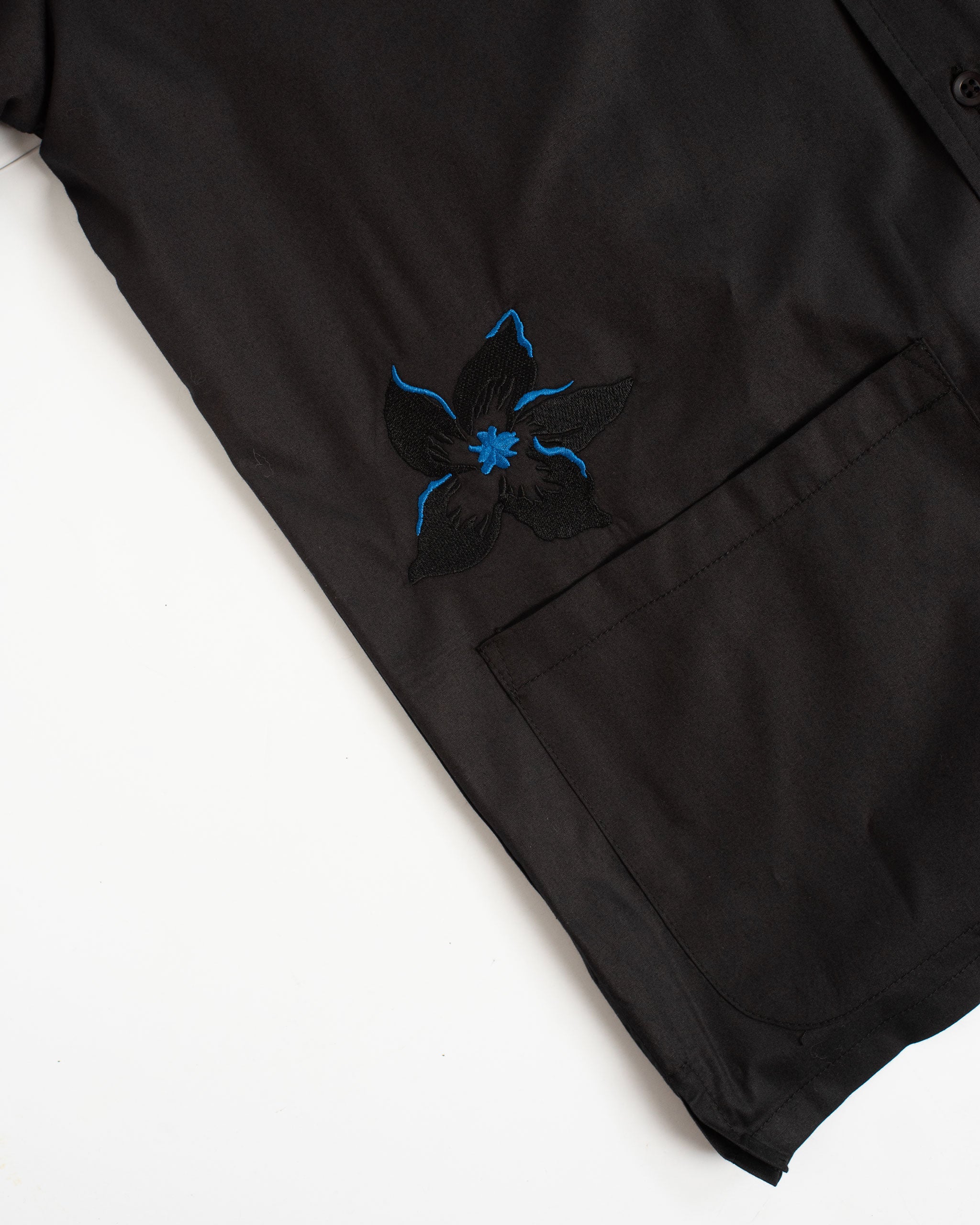 A close-up detail of the Bather Men's Black Embroidered Lily Camp Shirt, highlighting the intricate black lily embroidery with subtle blue accents and the front patch pocket.