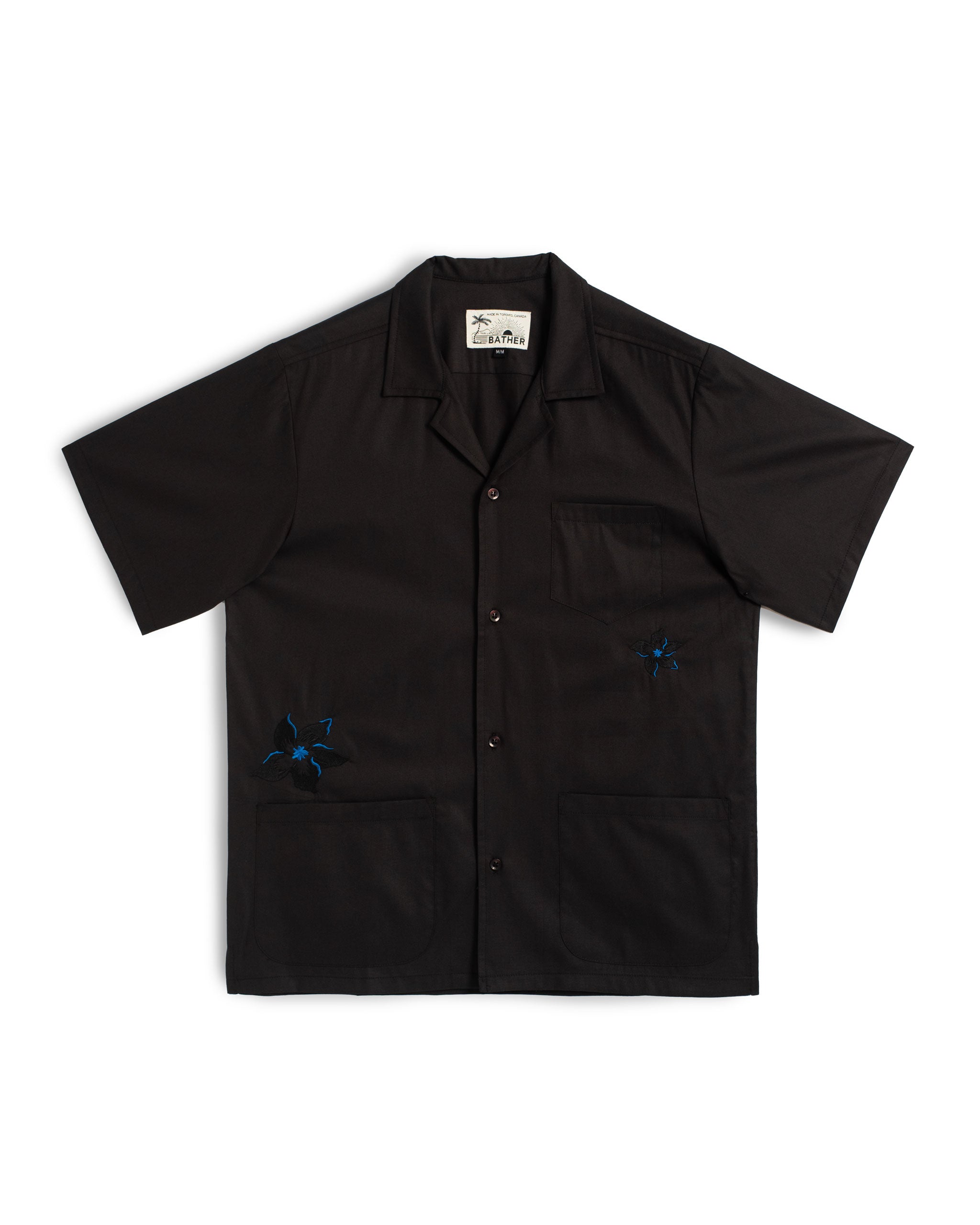 Front view of the Bather Men's Black Embroidered Lily Camp Shirt, featuring a black cotton-viscose blend fabric with white embroidered lily floral motifs, a single chest pocket, and a classic open camp collar against a white background.